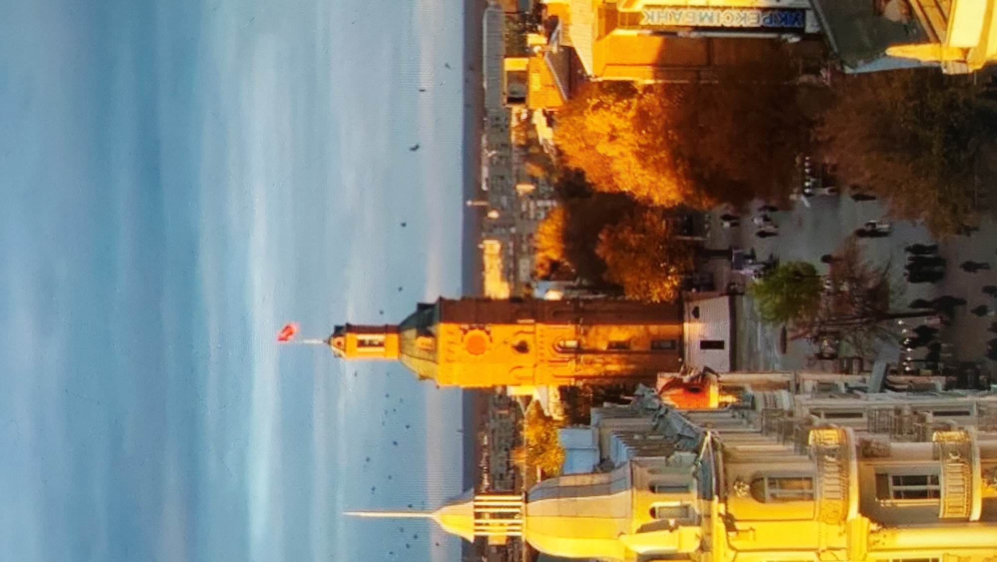 The image captures a city view from an elevated perspective during what appears to be late afternoon or early morning, given the warm, golden light illuminating the buildings and trees. Dominating the center of the frame is a tall, brick tower with a green conical roof, topped with a small flag. To the left of the tower, a lighter-colored building with a dome-like structure and a pointed spire is visible. The foreground shows the facades of ornate buildings with balconies, bathed in the golden sunlight. Below, a street is lined with trees displaying autumn foliage in vibrant yellows and oranges. Several people are scattered along the street, walking and gathered in small groups. The sky above is a clear blue with subtle horizontal cloud formations. The overall scene suggests an urban setting, possibly in a European city, with a mix of historic architecture and autumnal natural elements.