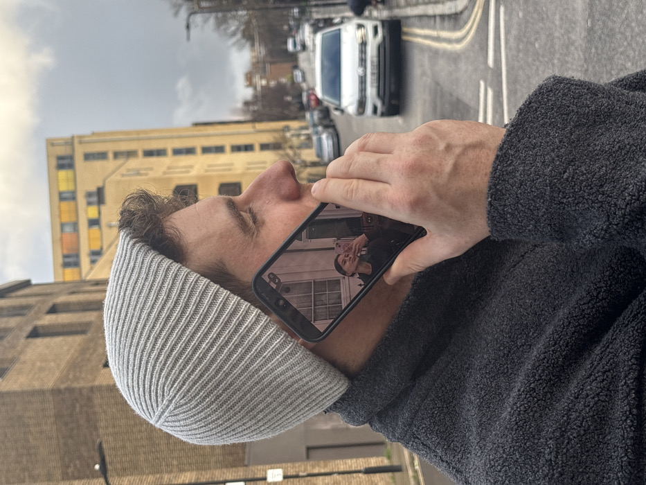 The image depicts an urban street scene in London, United Kingdom, under an overcast sky. In the foreground, a male, visible in profile from the chest up, faces right. He wears a light grey ribbed knit beanie and a dark grey textured fleece jacket. He holds a black smartphone to his right ear, appearing to be engaged in a call. The phone's screen displays a reflection or image of a female taking a selfie, holding a light pink phone to her face while making a "kiss" gesture. Her background appears to be an interior with a window and a door.
The background shows a paved street with double yellow lines, indicating no waiting or parking restrictions. Parked cars line the left side of the street. A white Volkswagen Tiguan SUV, displaying a UK license plate, is visible on the right side of the road, facing away from the viewer. Further down the street, more vehicles and buildings are present. On the left, a multi-story brick building stands, while in the mid-background, a taller, light-colored building features distinctive upper-floor windows with yellow, orange, and brown colored panels. Bare trees are visible along the street, suggesting a temperate climate or season.