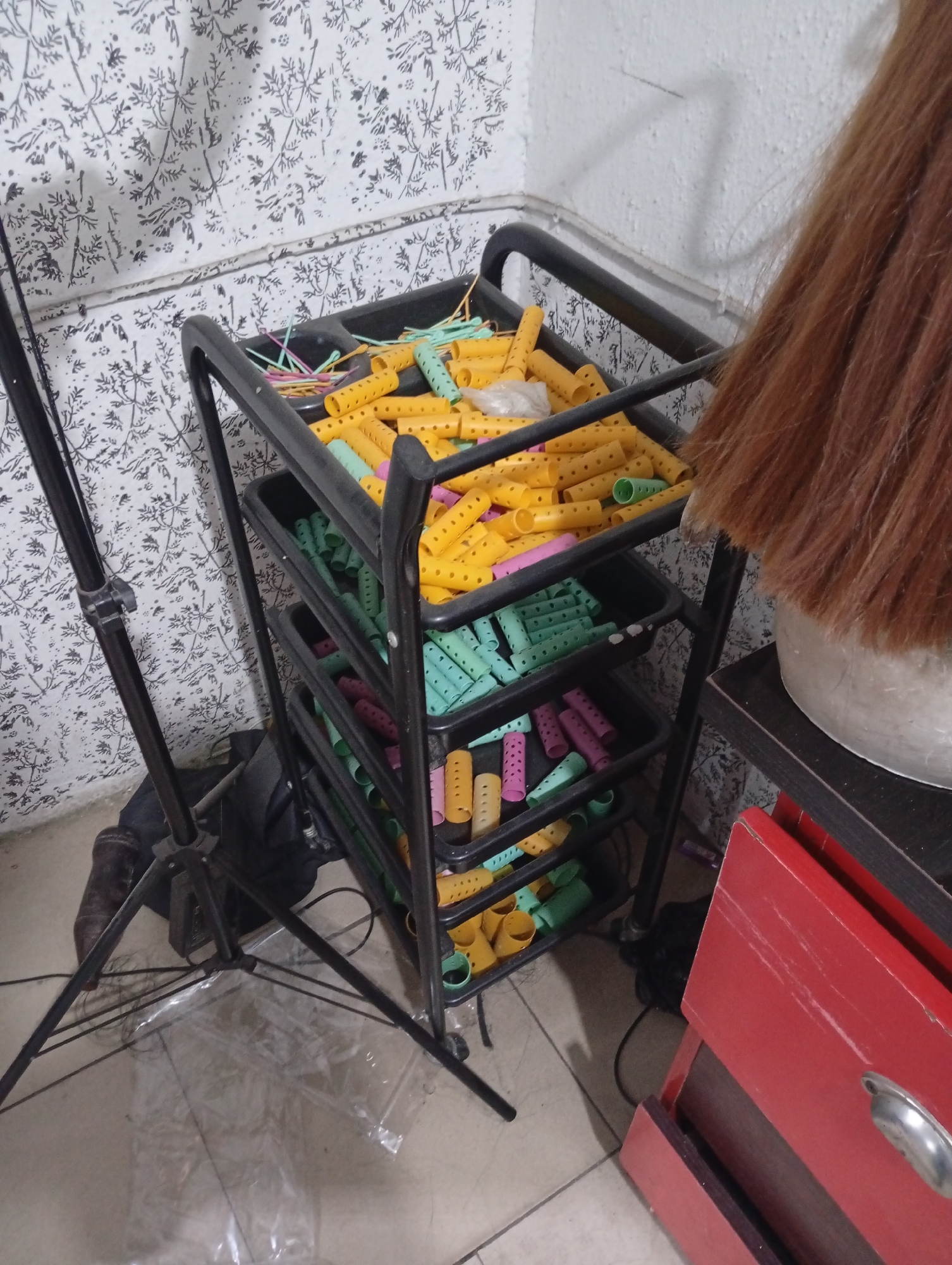 The image depicts a cluttered indoor setting, potentially a hair salon, in an unspecified location in Lokoja, Nigeria. A black metal rolling cart filled with various colored hair rollers (yellow, green, pink, and purple) stands against a wall with floral patterned wallpaper. A black camera tripod is visible to the left of the cart. To the right of the cart, part of a mannequin head with auburn-colored hair is seen, next to a red drawer unit. The floor is tiled. There are no people visible in the image.