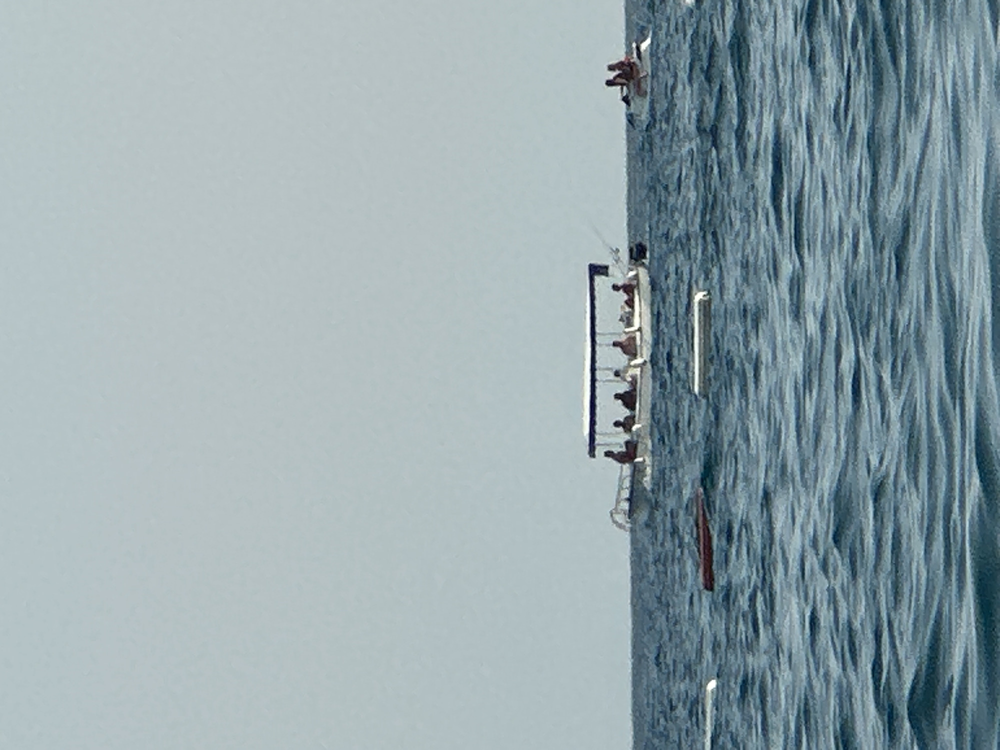 A wide shot captures a body of water under a pale blue sky. In the middle ground, a white boat with a canopy is traversing the choppy blue water. Six individuals are visible on the boat, some appearing to be seated. To the right of this boat, a smaller white boat carries three individuals. In the foreground, three white objects and one dark red object are floating on the water's surface. The scene is set at sea, near the coast of Boreti, Montenegro, indicated by the context of the image.