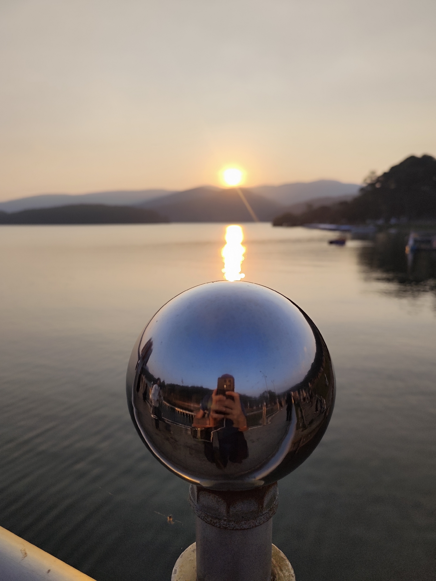 The image portrays a sunset over Xuan Huong Lake in Da Lat, Vietnam. A large, highly reflective chrome sphere, mounted as a finial on a weathered gray metal post, is centrally positioned in the foreground. The sphere's polished surface reflects the immediate surroundings behind the camera: a person is clearly visible holding a dark-colored smartphone, presumed to be the photographer, alongside distorted reflections of other indistinct human figures and architectural elements consistent with a lakeside promenade or pier.
Beyond the sphere, the main view encompasses the calm lake. The sun, a bright, intense orb, is low on the horizon, situated slightly to the left of the frame's center, casting a vivid, elongated vertical reflection across the water's gently rippled surface. Multiple layers of distant mountains or hills are silhouetted against the hazy sky, which transitions from a warm orange-yellow near the horizon to a lighter, muted beige overhead. The right side of the lake's shoreline features dark, indistinct forms, possibly vegetation or anchored boats.