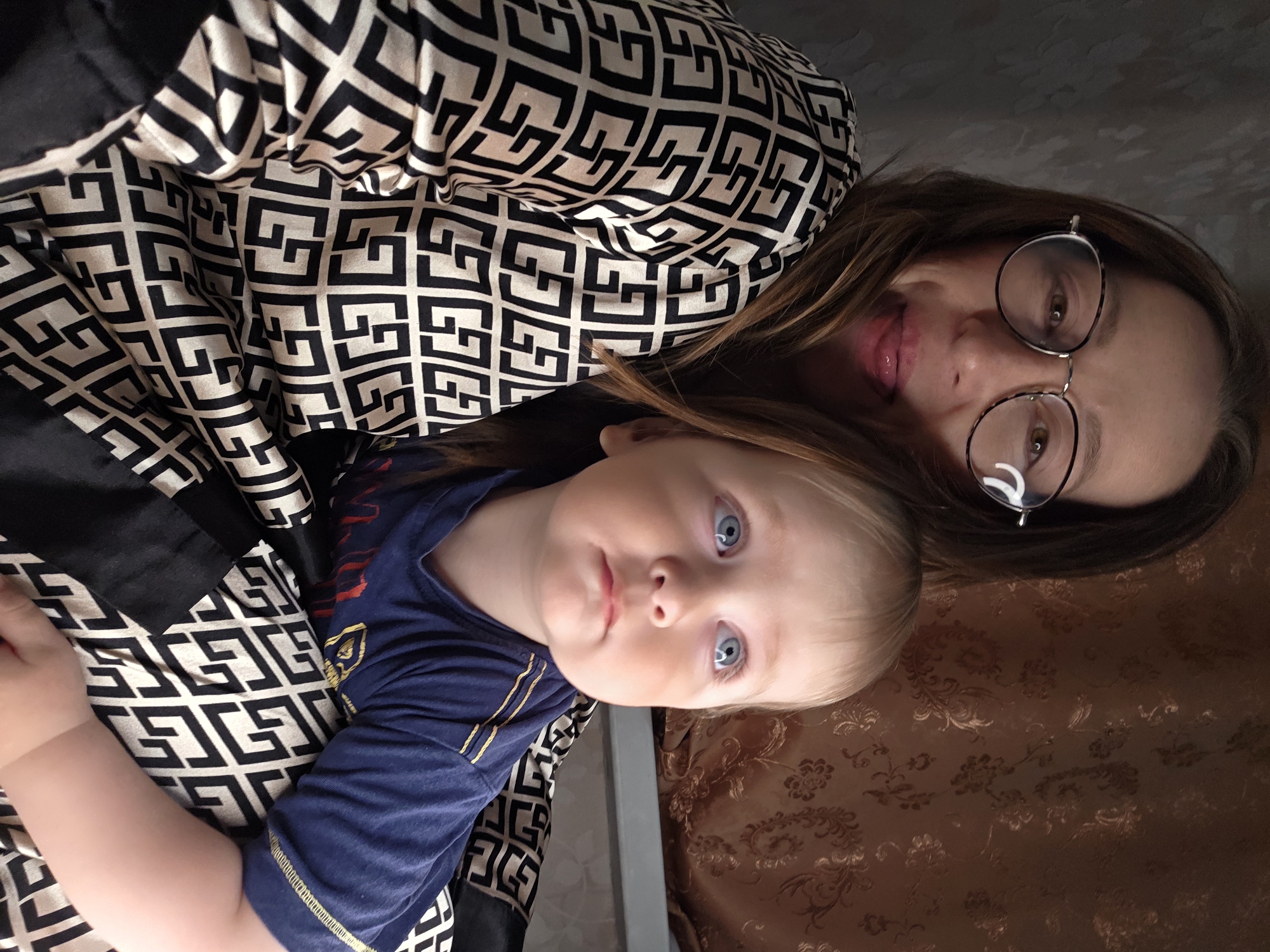 The image shows a person with light-brown hair, wearing glasses with a thin metal frame, and a geometric black and white patterned garment, posing with a baby in a blue t-shirt. The baby has light skin and blue eyes. The setting appears to be indoors, possibly on a bed or couch, as suggested by the brown patterned fabric in the background. While the city of Chaikovsky, Russia is provided, the image does not contain visual elements or clues to confirm the location.