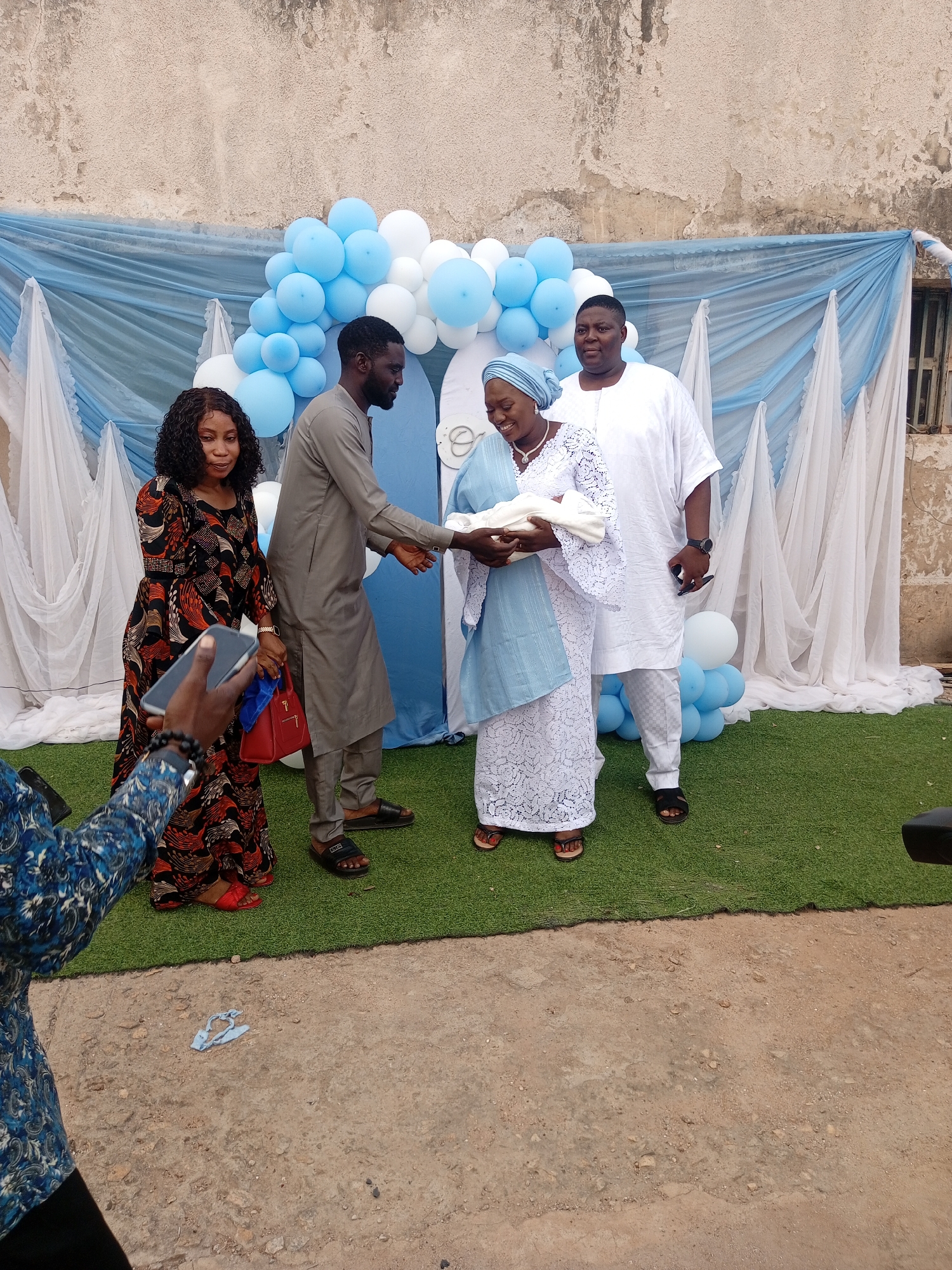 The image shows a group of four people standing outdoors, likely at a celebration event. A woman in a light blue head wrap and white lace dress holds a baby wrapped in a white cloth. A man in a grey outfit extends his hand toward the woman. Next to him stands a woman in a patterned dress holding a red bag, and on the right, a man in a white outfit stands with his hands at his sides. Behind them is a backdrop of light blue and white fabric draped around a white archway, decorated with blue and white balloons. Artificial turf covers the ground behind the people. The backdrop is positioned against a weathered wall with patchy paint and a small barred window on the right. A person holding a phone is partially visible on the left side of the image. The location appears to be in the city of Unknown, Unknown, but there are no clear indications of the city or specific location.