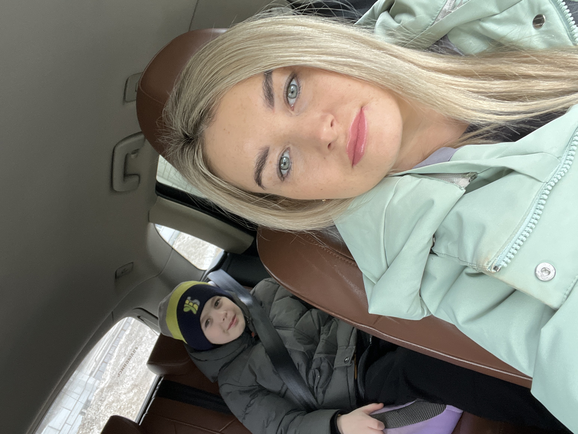 A medium, portrait-style shot from inside a car features a fair-skinned woman with blonde hair and blue-green eyes leaning towards the camera. She wears a light green jacket. Below her and to the left is a child in a gray winter jacket and a black and yellow striped beanie. The child is buckled into the brown leather car seat with a seatbelt. Outside the car window, a blurred view of a wintry streetscape is visible, suggesting an urban environment. Based on the available information and file metadata, the location is indicated as Chaikovsky, Russia.