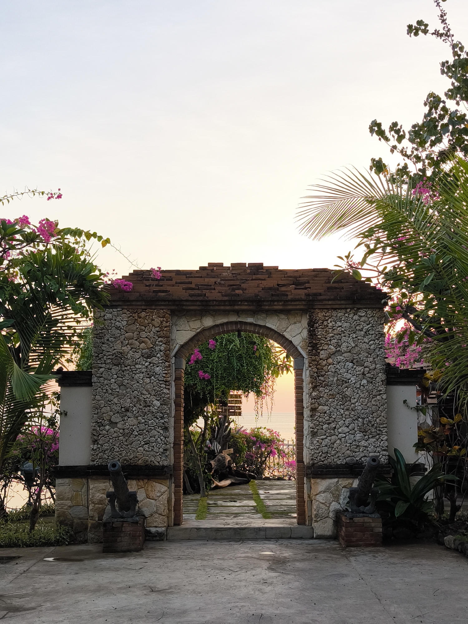 An arched stone gateway adorned with pink bougainvillea flowers leads to a sun-drenched vista of the ocean. The gateway is flanked by two antique cannons positioned on brick pedestals. Lush tropical foliage, including palm fronds, surrounds the entrance. The sky, a soft gradient of pink and orange, suggests either dawn or dusk. The ground in front of the gateway is paved with concrete, with some areas showing signs of wetness. The scene appears to be located in Labuan Bajo, Indonesia, possibly at a resort or private residence overlooking the sea. No people are visible in the image.