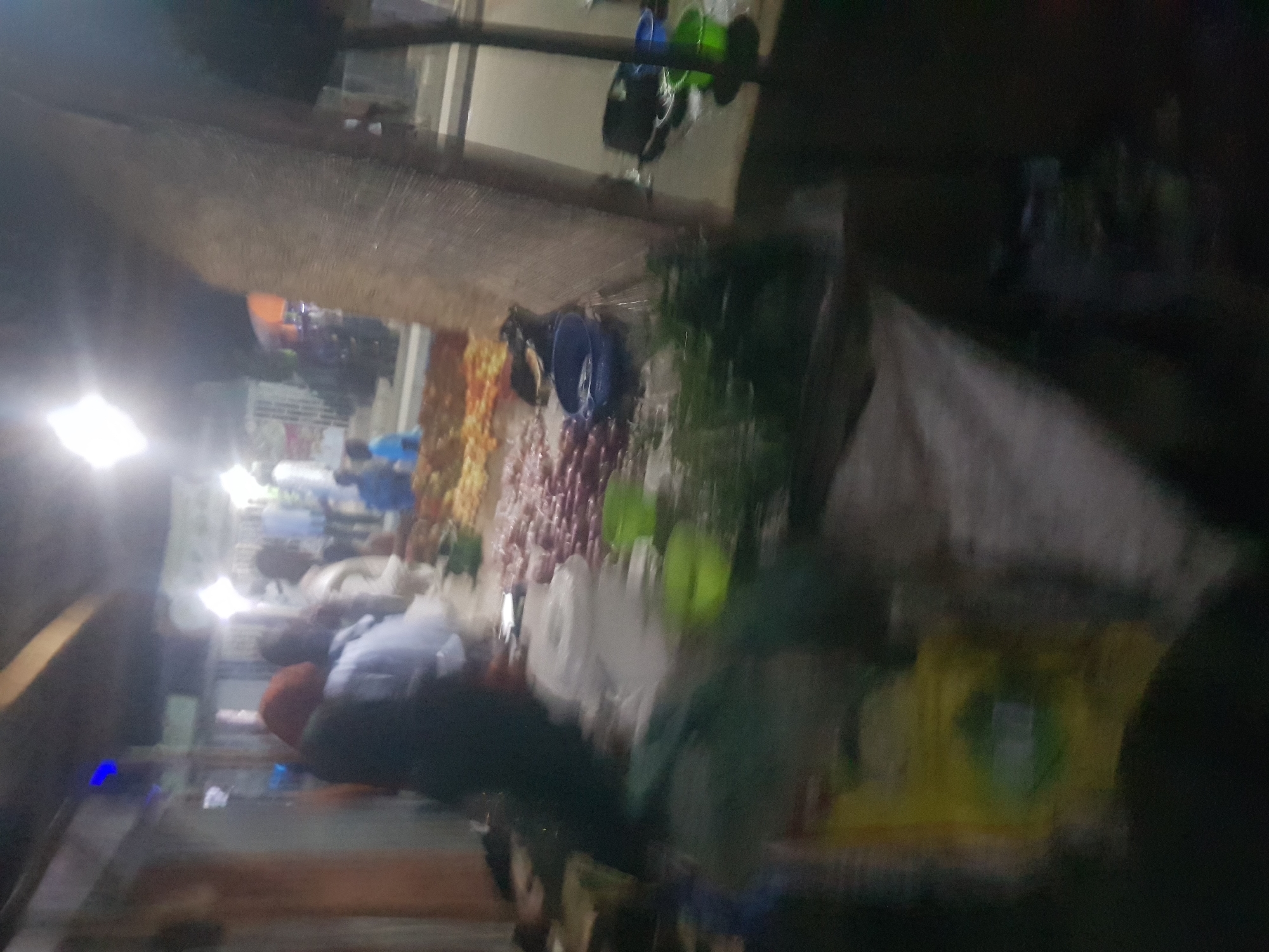 The image appears to show an outdoor market stall in Dabawa, Nigeria. People are visible, some wearing light-colored clothing and others wearing darker clothing, suggesting a variety of customers and vendors. The stall itself displays a range of goods, including produce such as oranges, onions, and leafy greens, likely for sale. Other items, possibly cookware or other household goods, are also visible. The lighting is somewhat dim.