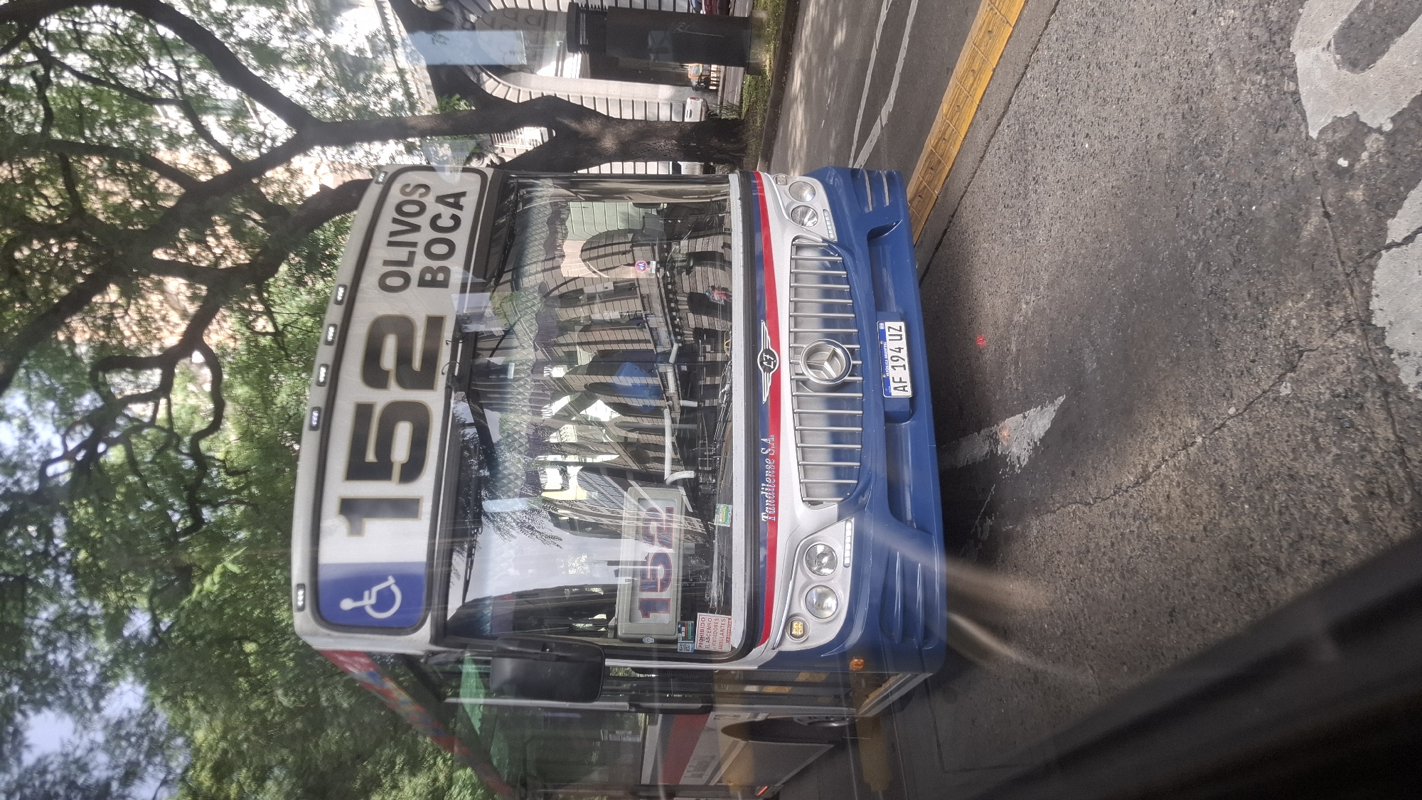 The image shows the front of a blue and white bus, route number 152, with "OLIVOS BOCA" displayed above the number. The license plate reads "AF 194 UZ." The bus has a handicap symbol on the top left corner. The bus appears to be in a city environment, likely in Buenos Aires, Argentina. A tree is visible on the left side. Part of the street is seen on the right. There is text reading "Tandilense S.A." on the front of the bus. The image is taken through a window, possibly of another vehicle.