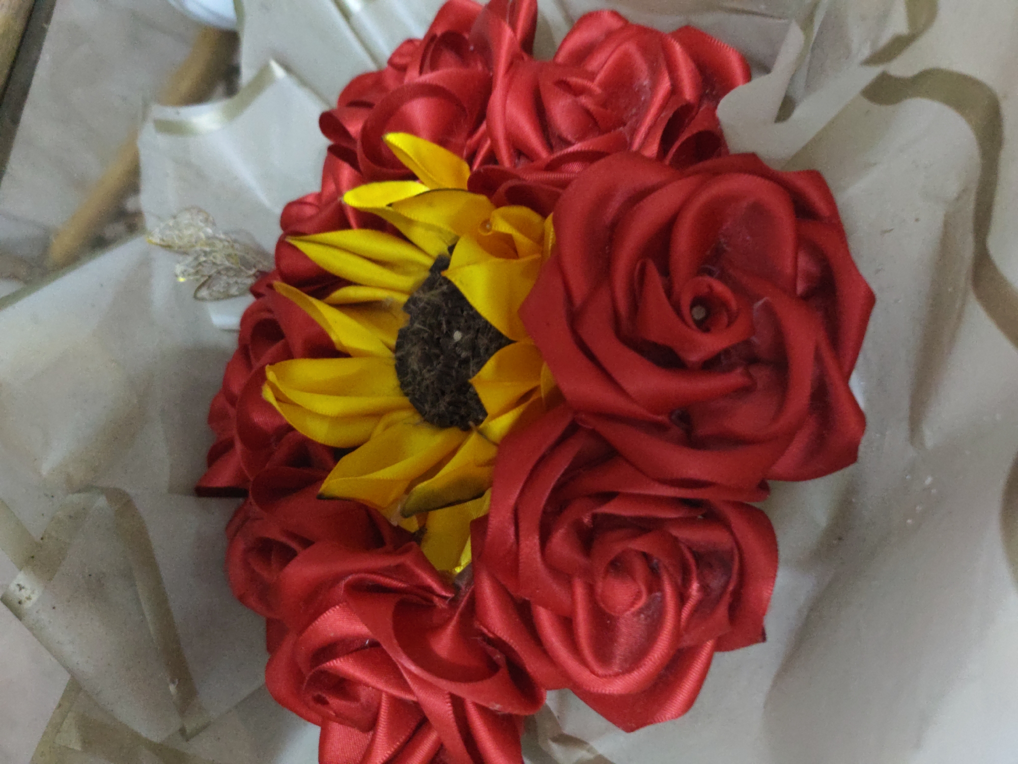 A bouquet of red ribbon roses and a single yellow ribbon sunflower is presented on a light-colored, textured surface that appears to be some kind of fabric or paper wrap, possibly part of a gift. The bouquet is arranged in a cluster, with the sunflower situated slightly off-center amidst the vibrant red roses. The ribbon roses are tightly coiled to mimic natural rose petals, and the sunflower is similarly constructed from fabric petals surrounding a dark center. A delicate, shimmering embellishment, possibly a small butterfly or insect made of wire or foil, is positioned to the left of the bouquet, partially obscured by the flowers. The background is softly blurred, suggesting an indoor setting or a focus on the bouquet itself. The scene does not provide any specific visual cues to indicate a location in Piloncito, Venezuela.