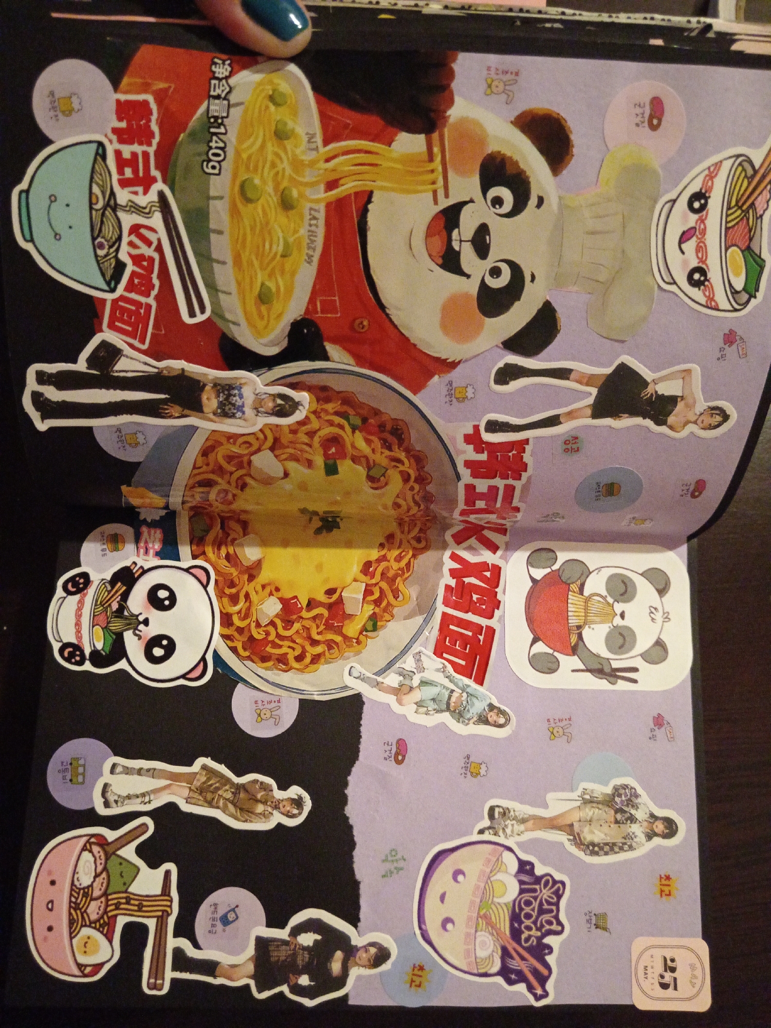 This is a close-up photograph of an open scrapbook, likely a journal or art project. The left page is black, while the right page has a light purple background. The scrapbook is filled with various stickers, images, and text related to Korean and Chinese instant noodles.
A large image of a panda bear character associated with noodles is prominently featured on the upper portion of the left page. Various cartoon-style stickers of bowls of noodles, pandas eating noodles, and Korean/Chinese text are scattered around the page. Additionally, there are several stickers featuring images of women, seemingly fashion or model shots.
The layout is collage-like, with overlapping stickers and images. The style of the scrapbook suggests a playful, artistic expression, possibly themed around Asian cuisine and culture. The presence of Korean and Chinese text and noodle depictions emphasizes this theme.