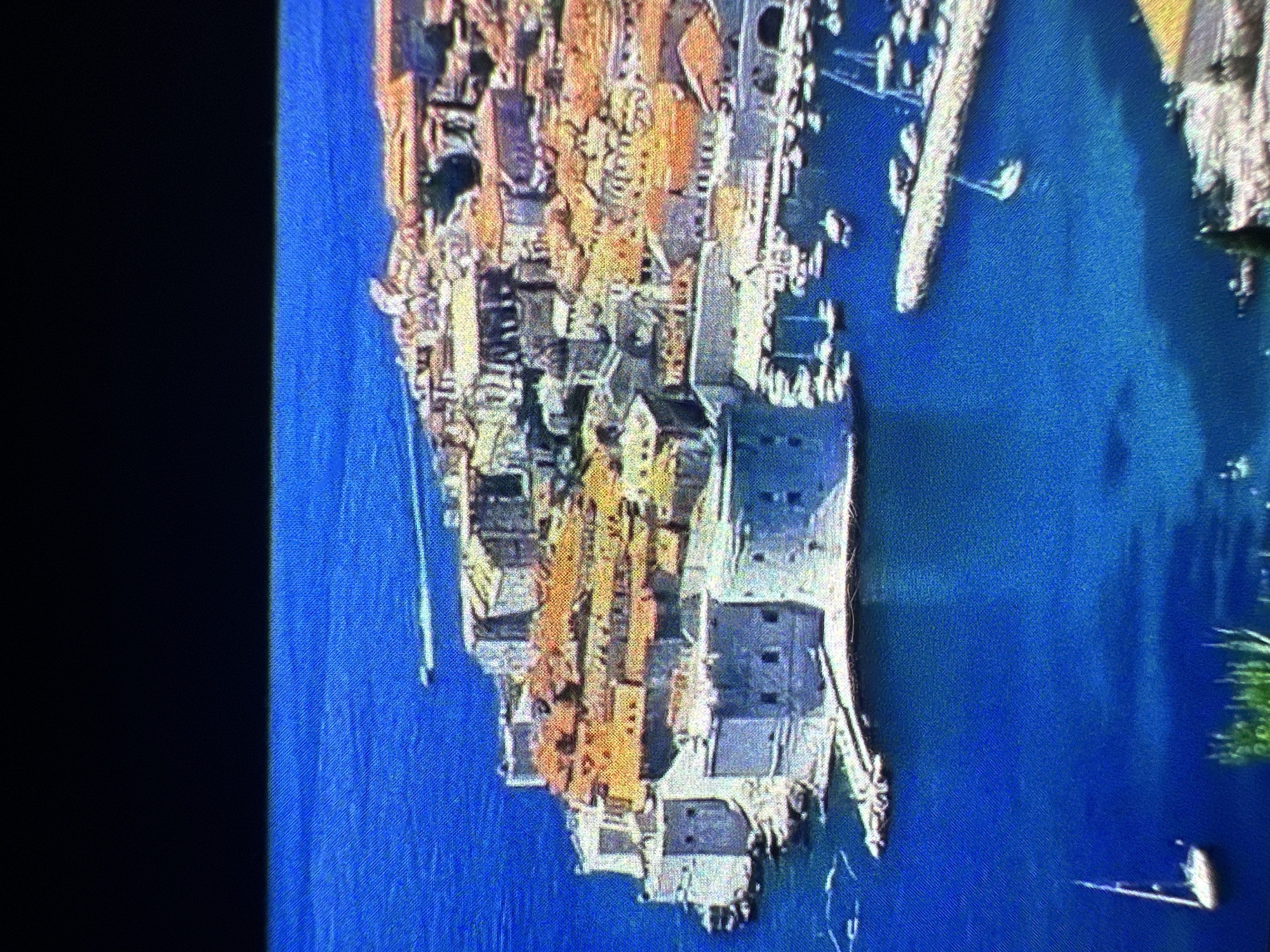 The image shows an aerial view of a fortified city on a peninsula jutting into a bright blue sea. The city has numerous buildings with orange tiled roofs clustered together. Large, stone fortifications with rounded towers surround the seaward side of the city, with ramparts and defensive structures visible. A harbor is located within the city walls, filled with numerous small boats and yachts. A breakwater extends from the city into the sea, with more boats moored alongside it. In the foreground, a single white sailboat with its mast up is visible in the open water, and a second, smaller boat is near the shore on the left. The water is calm and clear, reflecting the blue of the sky. A dark, horizontal band stretches across the top of the image, obscuring the upper portion of the scene.