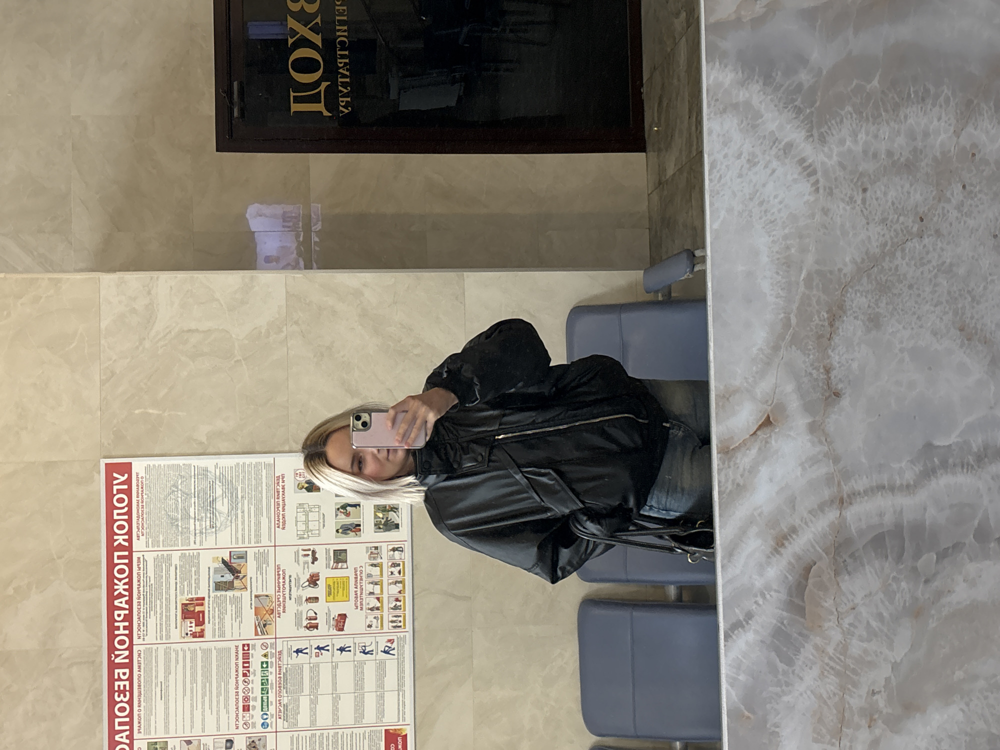 A woman is taking a selfie in front of a wall-mounted fire safety evacuation plan written in Russian, which includes safety regulations and diagrams. The wall is marbled. In the top center of the image is a dark brown door with the word "Вход" written in gold lettering, which translates to "Entrance". The reflection indicates the scene may be indoors. The woman is wearing a dark jacket and holding a pink phone in front of her face. There are rows of blue chairs where she is sitting. There are hints of the city name Saint Petersburg, Russia based on the text found on the sign.