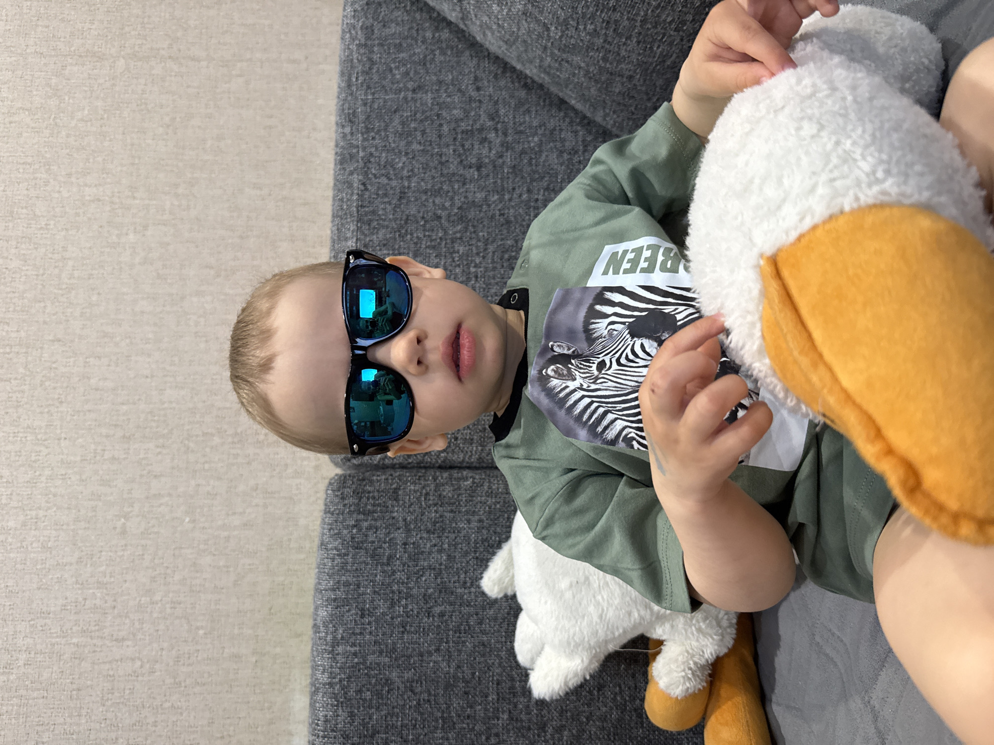 A small child wearing blue-mirrored sunglasses, a green zebra-print t-shirt, and green shorts is sitting on a gray couch. The child is holding a large white and yellow stuffed duck. The location is Rusanovo, Russia.