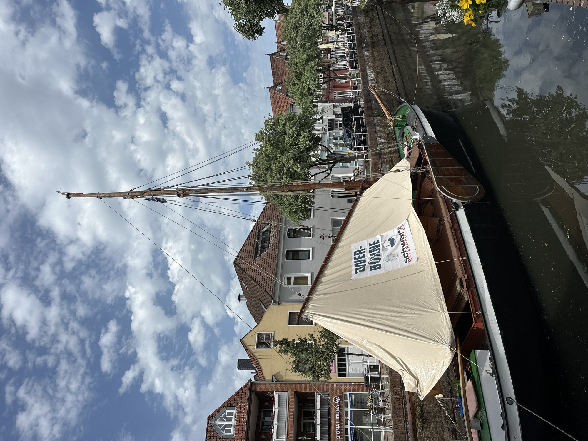 A sunlit, outdoor scene features a historic sailing boat moored on a narrow canal in the city of Buxtehude, Germany. The boat is rigged with a tall, wooden mast and a beige-colored sail. A promotional banner is affixed to the sail. Tightly packed buildings line the canal's edge. Their architectural styles vary, featuring red tile roofs and a mix of brick and plaster façades. Several trees are interspersed along the canal, partially obscuring the building fronts. The sky is bright blue, filled with scattered white clouds. The dark water of the canal reflects the buildings and trees.