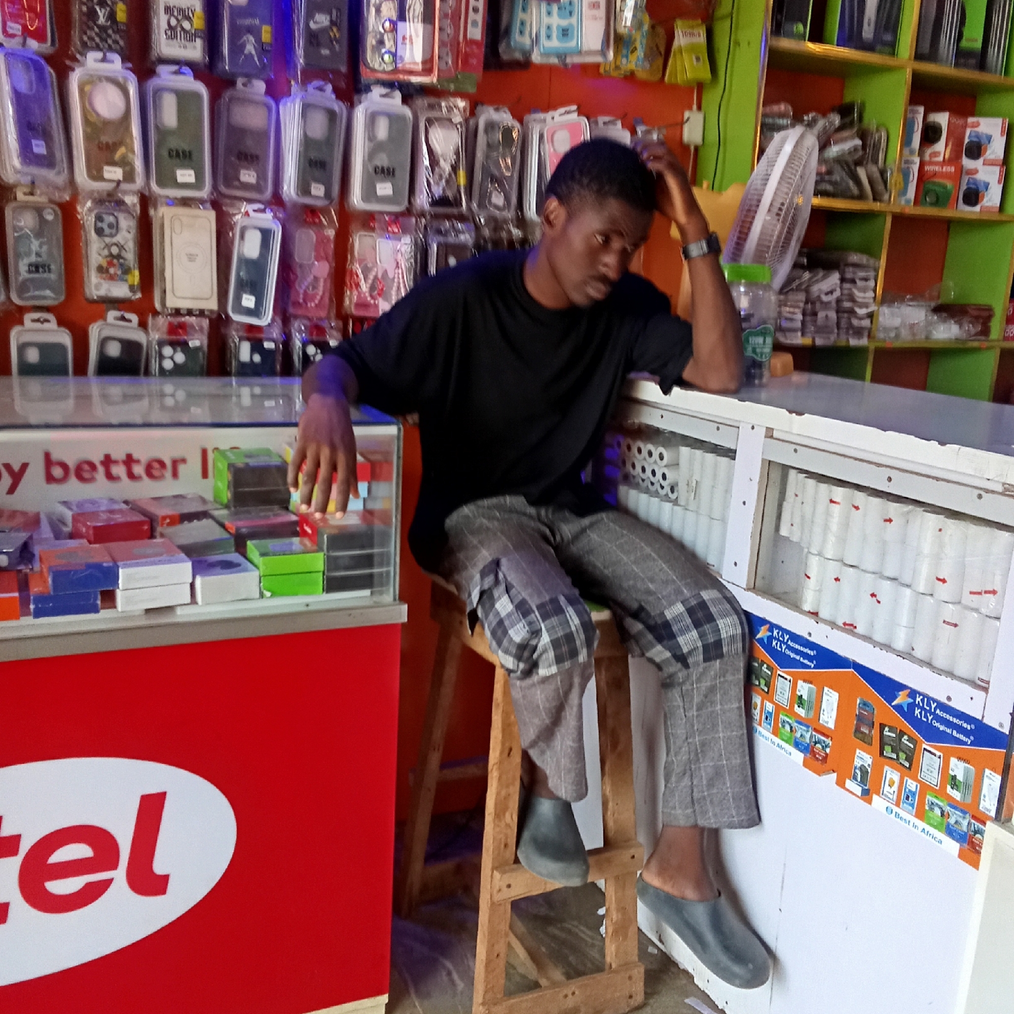 A young man is sitting on a wooden stool behind a counter in a retail shop in Akwanga, Nigeria. The store sells phone accessories and electronic devices. The man is wearing a black t-shirt and gray plaid pants. He has one hand resting on his head. The counter is lined with a display case containing small boxed items and rolls of paper. Behind the counter, there are shelves filled with various products, including phone cases. A fan is also visible behind the man. The exterior of the counter features the branding of the telecommunications company Airtel and the battery brand KLY.