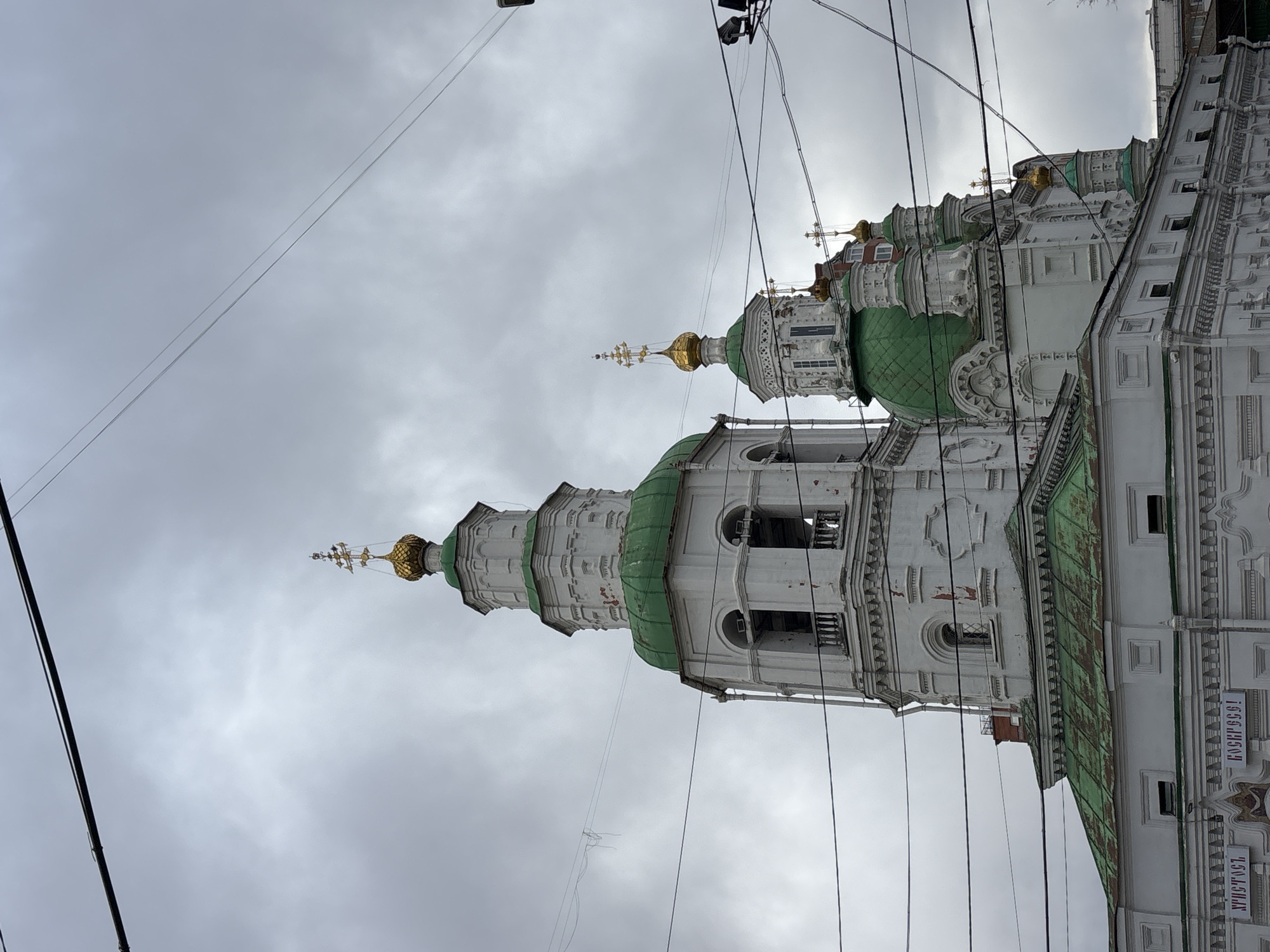 The image depicts the Annunciation Cathedral in Krasnoyarsk, Russia, viewed from a low angle. The cathedral's white facade, adorned with elaborate detailing, is topped by green domes and golden crosses. Overhead power lines crisscross the sky, which is overcast with gray clouds. The building features multiple tiers and arches, characteristic of Russian Orthodox architecture. A sign with lettering in Cyrillic script is visible on the side of the building.