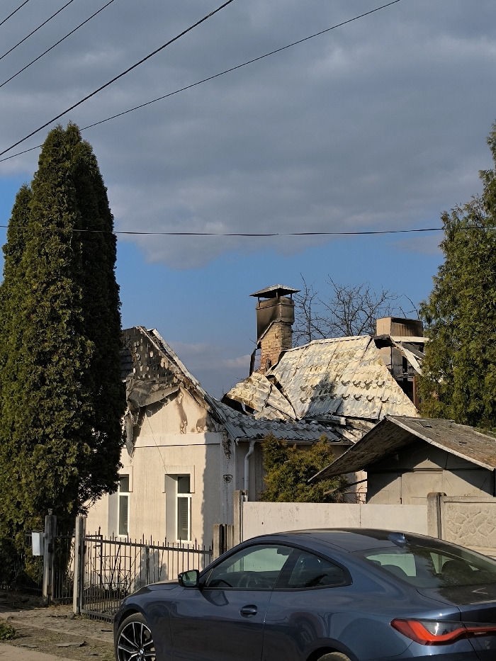 The image displays a residential street in Vinnytsia, Ukraine, featuring two heavily damaged houses under a partly cloudy sky, with a modern vehicle parked in the foreground.
In the midground, the primary structure on the left exhibits a partially collapsed and charred roof, with exposed blackened timbers and soot stains visible on the upper section of its light-colored facade. Two rectangular windows are visible on the lower portion of this house. Adjacent to it on the right, a second house shows a severely buckled, torn, and collapsed metal-sheet roof. A blackened brick chimney with a capped top rises above this damaged roof. Behind the structures, bare deciduous tree branches are visible, interspersed with evergreen foliage.
A tall, slender evergreen tree, likely a cypress or thuja, stands prominently on the left side of the frame, partially obscuring the left house. Another evergreen tree is visible on the far right. Multiple overhead utility lines traverse the upper portion of the sky.
In the foreground, on an unpaved or dirt road, the rear section of a dark blue two-door coupe car is parked, facing right. The vehicle appears intact and undamaged. A low concrete or stone wall separates the properties from the unpaved road. No individuals are visible in the scene.