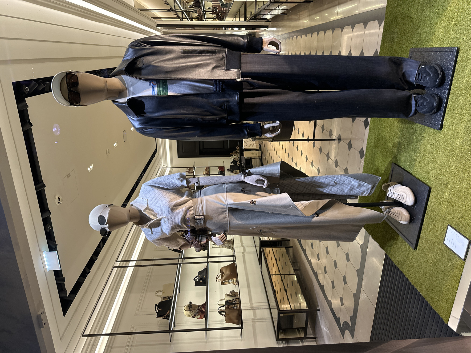 A retail store window display features two mannequins dressed in fashion apparel. The mannequin on the left is female, wearing a light-colored, collared shirt and a long, flowing skirt. It is accessorized with sunglasses and a handbag. The mannequin on the right is male, dressed in a dark leather jacket over a graphic t-shirt and dark pinstripe pants, also wearing sunglasses. Both mannequins stand on a green turf-like surface, in front of shelving units displaying handbags and other accessories. The store interior is visible behind the mannequins, with checkered flooring and recessed lighting. This display is located in a retail environment in Vienna, Austria.