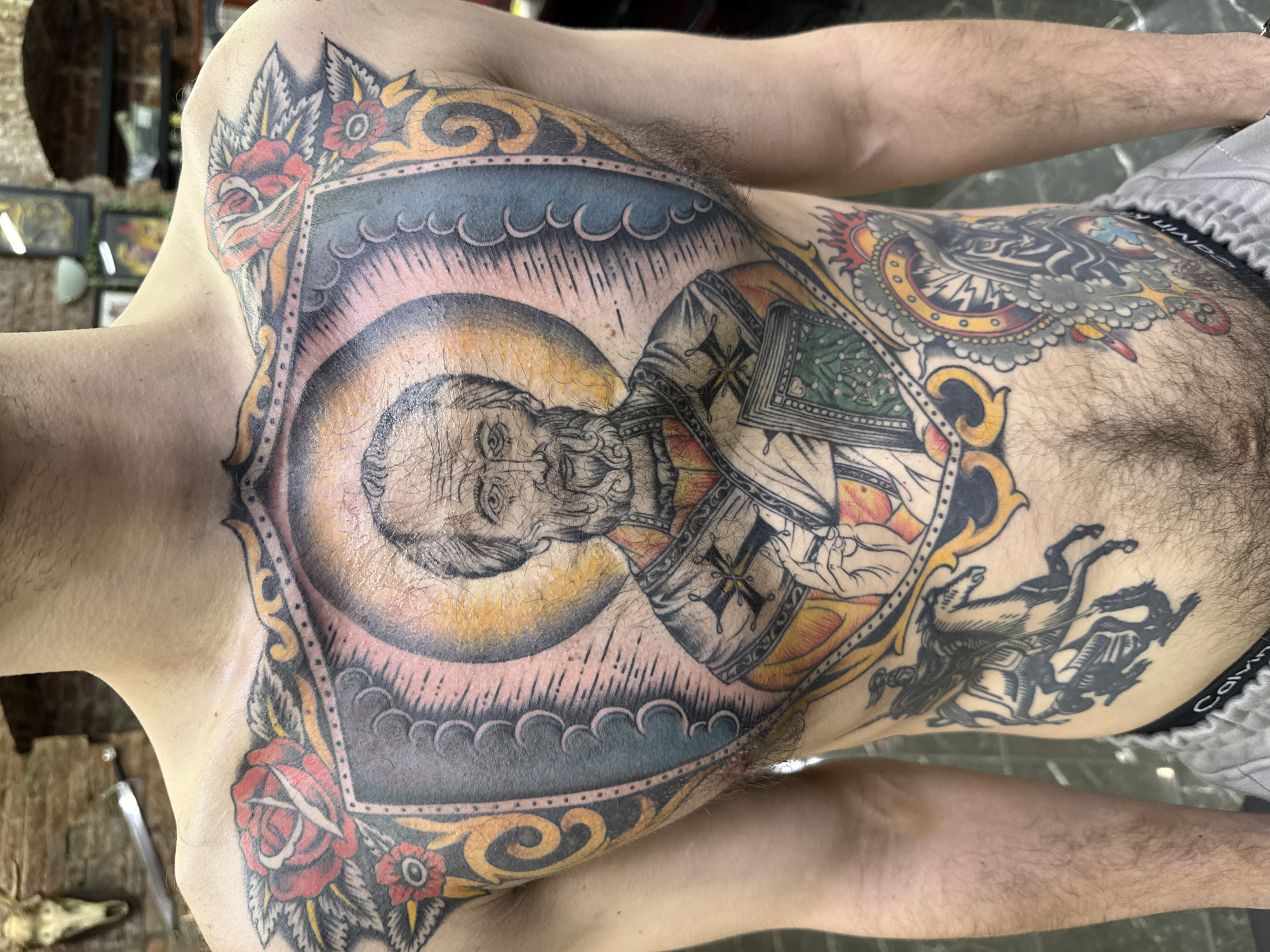 A close-up, chest-up shot of a person with extensive tattoos. The main tattoo covers the chest, depicting a framed religious figure with a halo. The frame is decorated with roses and ornate designs. Additional tattoos are visible on the upper abdomen, including a horse-like figure. The person is wearing grey underwear. The background is out of focus but appears to be an interior space with a wooden table and what looks like a sword and skull-like object. The location (Moscow, Russia) is not discernible from the image.