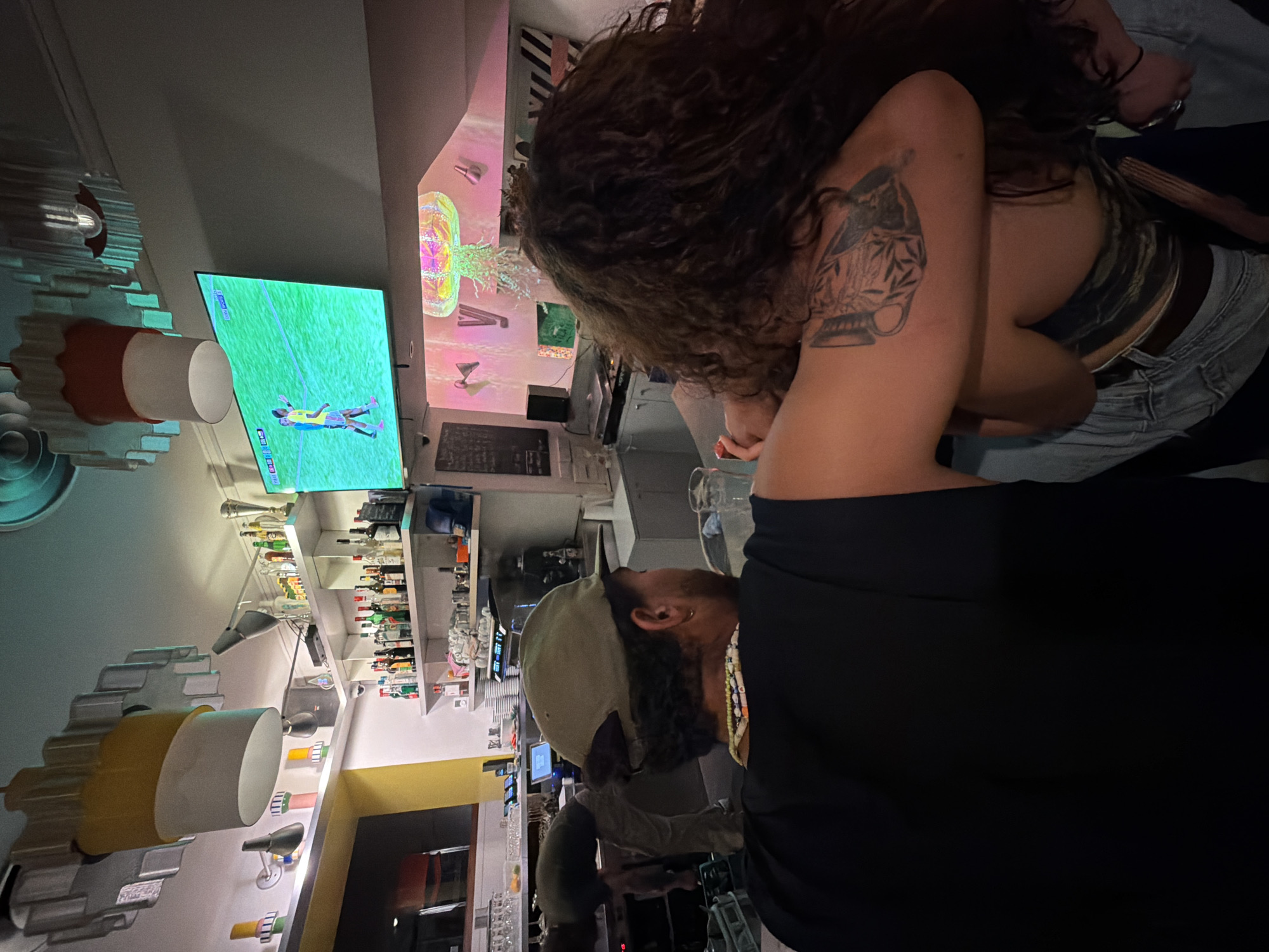 In a dimly lit establishment, likely a bar or restaurant, two individuals are captured from behind, engaged in an embrace. The person in the foreground, facing away from the camera, wears a black t-shirt and a green baseball cap. A beaded necklace adorns their neck. Their left arm is around the shoulders of the second individual, whose long, curly brown hair cascades down their back. This person is wearing a patterned top and blue jeans, and a tattoo is visible on their left arm.
Above them, a large television screen displays a live soccer match. The background reveals shelves stocked with various bottles, suggesting a bar area. The ceiling features decorative elements, one resembling a large, stylized gear. The overall ambiance is casual and intimate, with a focus on shared activity. The establishment appears to be in Lyon, France, given the context.