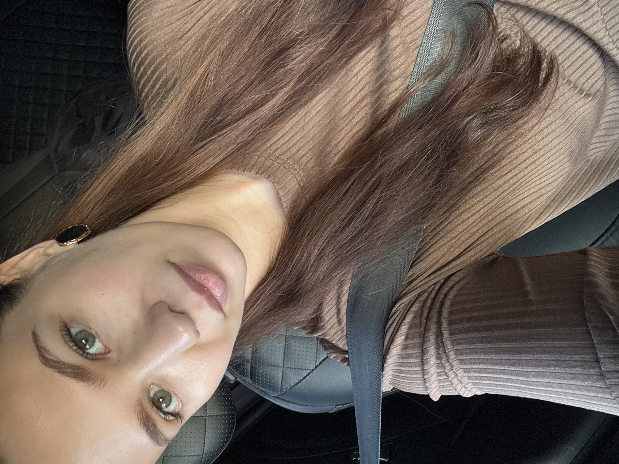 A close-up portrait shows a young woman inside a car in Chaikovsky, Russia, taken from a slightly upward, rotated angle, suggesting a selfie. The woman occupies the majority of the frame, with her head and upper torso visible. She has light skin, striking green or hazel eyes with well-defined dark eyebrows, and light pink lips. Her long, straight brown hair flows over her shoulders and chest, partly covering her clothing.
She is wearing a form-fitting, long-sleeved top made of ribbed, light brown or nude-colored fabric, which is visible across her chest and arms. A dark seatbelt diagonally crosses her torso, from her left shoulder down towards the right. On her visible left ear, she wears a small earring with a dark center and a golden rim.
The background reveals parts of the car's interior, specifically a dark, possibly black, leather car seat with a quilted pattern visible on the headrest and backrest. The lighting appears soft and natural, highlighting her features without harsh shadows. Her expression is neutral, with her lips slightly parted, and she looks directly towards the camera. There is no visible text in the image.