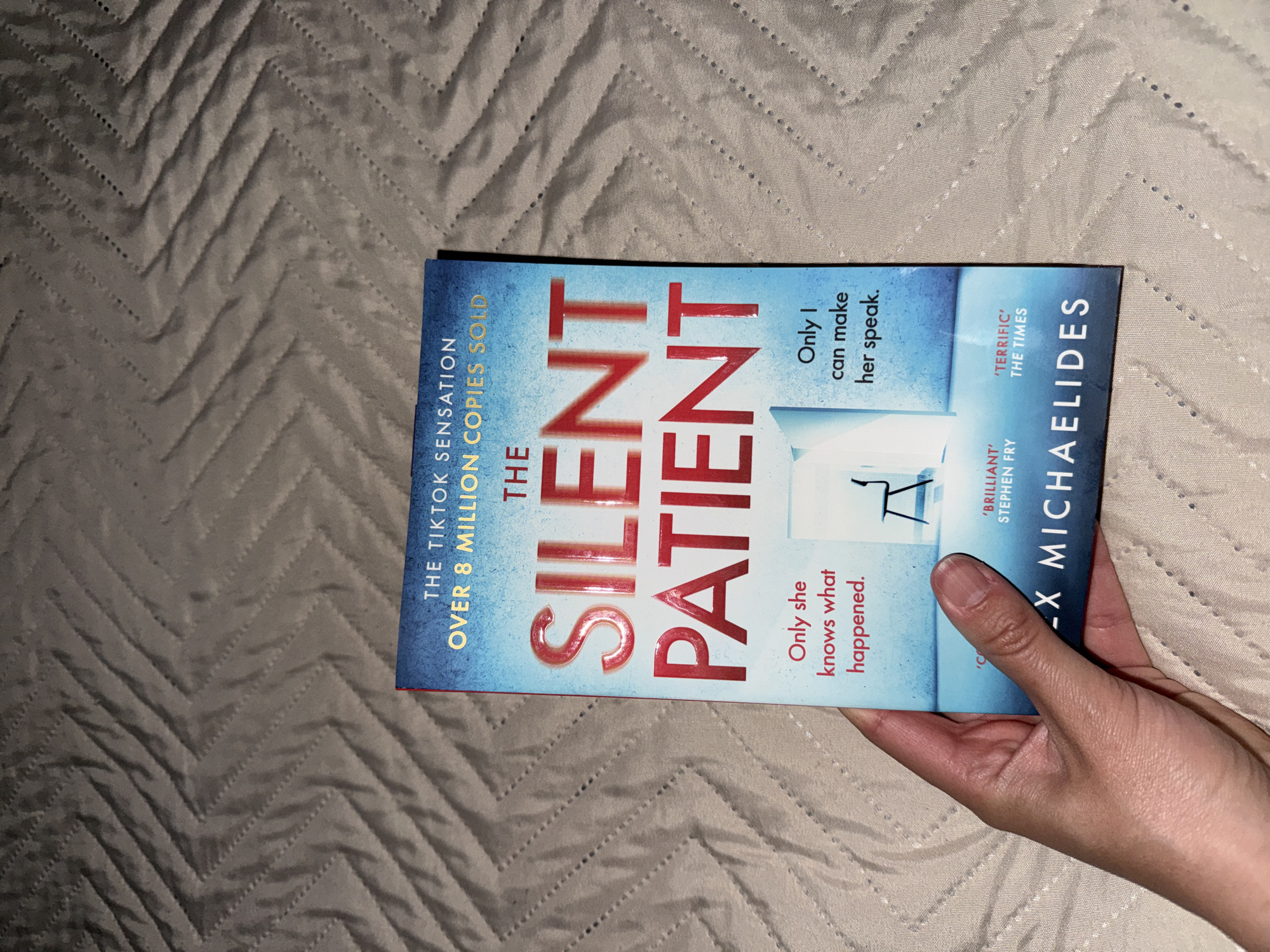 A slightly rotated, close-up shot shows a light-skinned hand holding a book against a background of a gray quilted blanket. The blanket features a prominent chevron or zigzag stitched pattern. The book, titled "THE SILENT PATIENT" by Alex Michaelides, is held by the right hand, with fingers curled around the bottom and thumb resting along the right edge.
The book cover has a gradient design, transitioning from a darker blue at the top and bottom to a lighter, almost white, blue in the middle. The title "THE SILENT PATIENT" is written in large, bold red letters. A graphic of a white, sparsely furnished room with a single chair is visible in the lighter blue section of the cover.
Several promotional texts are visible on the cover:
* "THE TIKTOK SENSATION"
* "OVER 8 MILLION COPIES SOLD"
* "Only she knows what happened."
* "Only I can make her speak."
* "'BRILLIANT' STEPHEN FRY"
* "'TERRIFIC' THE TIMES"
The lighting suggests an indoor setting, possibly at night or in a dimly lit room, with artificial light illuminating the book and hand. The hand appears to have natural, unpolished nails. The overall impression is that of someone preparing to read or having just finished reading the book.