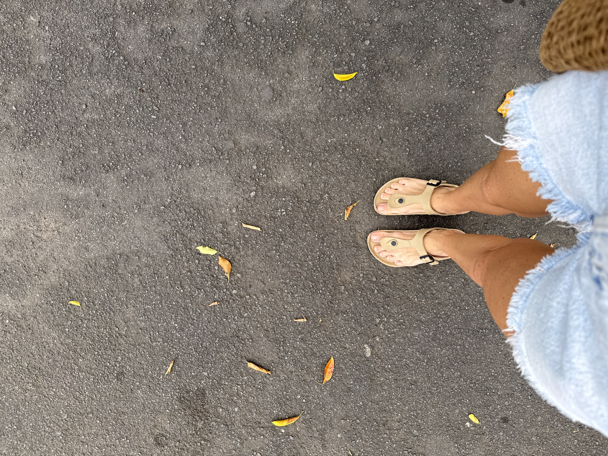 The image presents an overhead view looking down at a person's lower body and the ground. The scene is set on a dark grey asphalt surface, characterized by a coarse, textured appearance of small pebbles and aggregate. Scattered across this ground are several fallen leaves, predominantly in shades of yellow and light brown, suggesting a natural outdoor environment.
In the right portion of the frame, the person's legs and feet are visible. They are wearing light-colored, possibly beige or tan, open-toed sandals with a T-strap design and a buckle detail. Their bare feet are visible within the sandals, and their skin appears tanned. The person is dressed in light blue denim shorts, notable for their frayed or distressed hems, indicating a casual style. A small part of what might be a straw hat is visible at the very top right edge of the image.
The lighting suggests it is daytime. The attire – sandals and denim shorts – points to a warm climate, consistent with the location context of Kuta Selatan, Indonesia. There are no discernible activities beyond standing or taking a step, and no visible text is present in the image.