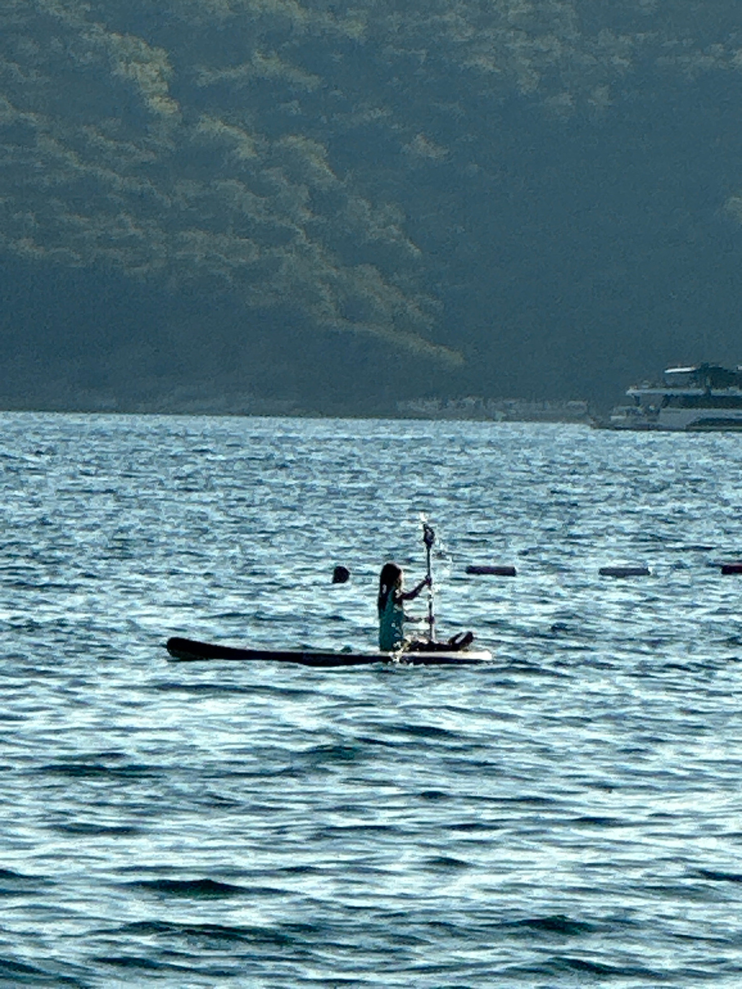 A person is paddleboarding on a body of water. The person is sitting on the paddleboard, holding a paddle. The water is choppy, and there is a boat in the background. The background also shows a dark, tree-covered hill. The image is taken from a distance, with the paddleboarder in the center of the frame. The sky is overcast, and the lighting is dim. The image appears to be taken on a lake or a calm sea. The location is identified as Boreti, Montenegro.