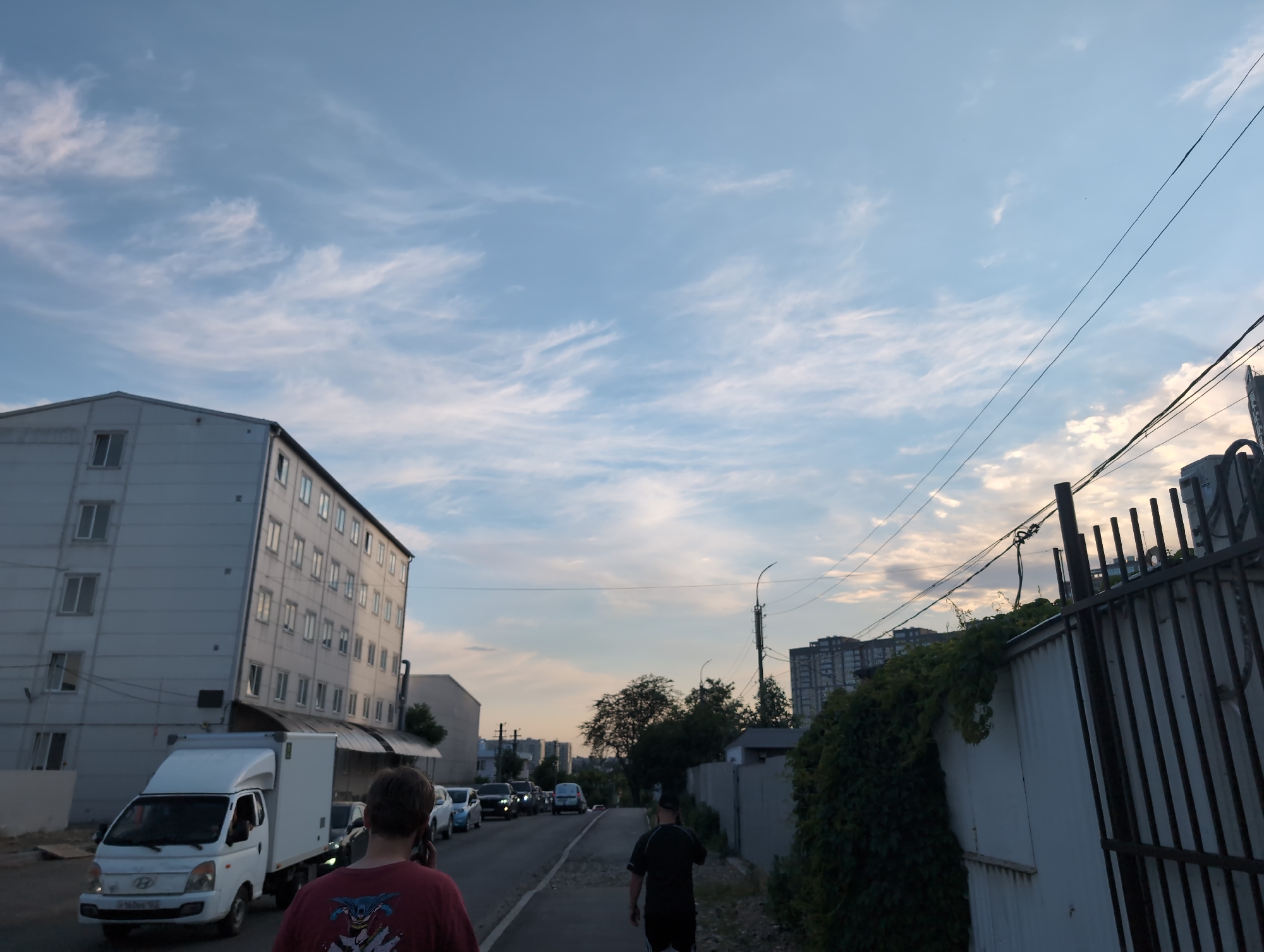 The image depicts a street scene in Krasnodar, Russia, during what appears to be late afternoon or early evening, with a cloudy sky transitioning to dusk. In the foreground, a person wearing a red shirt with a graphic print is walking away from the camera, seemingly engaged in a phone conversation. To the left, a white box truck is parked on the side of the road, with its driver's side door open. Further down the street, a line of parked cars is visible.
Dominating the left side of the image is a multi-story building with a light-colored facade and numerous windows. On the right side, a dark metal fence runs along the edge of the property, partially overgrown with green vines. In the background, several other buildings, including taller apartment complexes, are visible. A street light on a pole stands near the fence. The overall environment suggests an urban setting with a mix of residential and commercial structures.