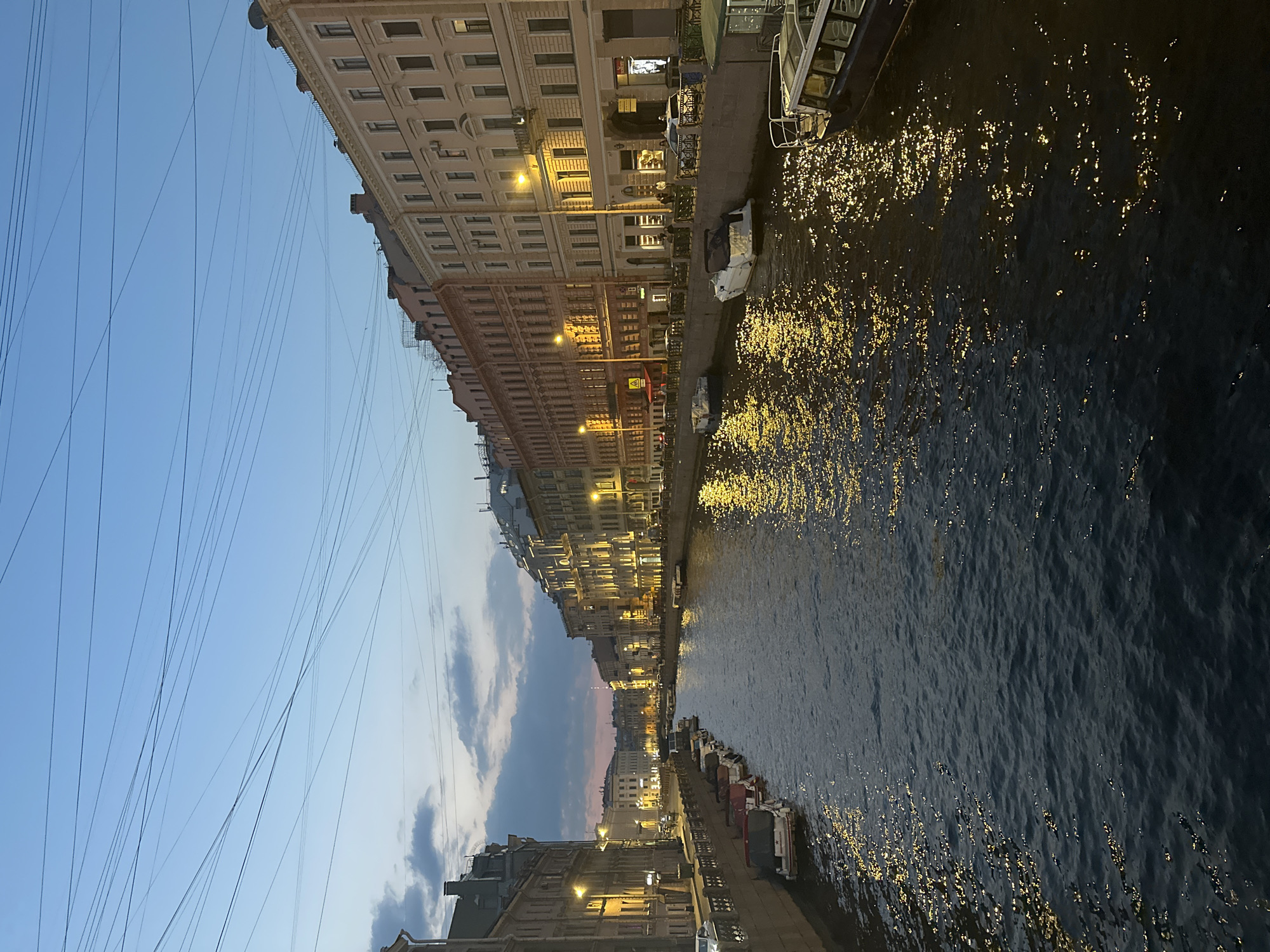 The image captures a canal scene at dusk in what appears to be an urban setting. On either side of the canal are buildings that appear to be of historical architecture, illuminated by streetlights. Boats are docked along the canal sides. Above, the sky is a mix of blue and dusk colors. There are many utility or electrical lines visible against the sky. The water in the canal is dark, with light reflecting on the surface.