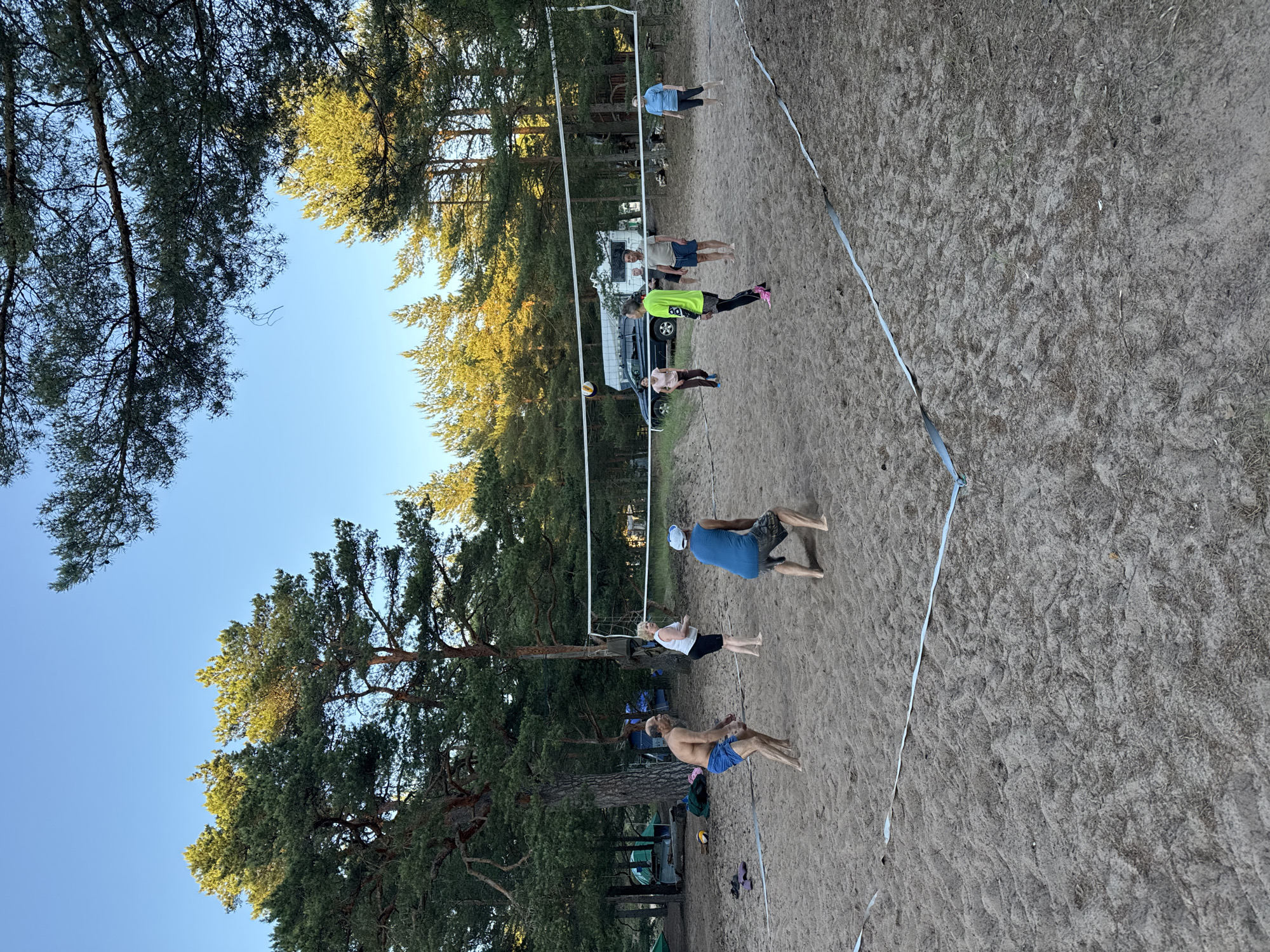 The image depicts a beach volleyball court set up on a sandy area, surrounded by tall pine trees. The sky is clear and blue. Several people are actively playing volleyball. On one side of the net, a man in blue shorts is poised to hit the ball, while a woman in a white tank top and black shorts stands nearby. On the other side, a man in blue shorts and a blue tank top is in motion, with a volleyball in the air above the net. Another person in a bright green shirt and black leggings is also on this side, facing the net. A woman in a light blue shirt and dark pants is positioned further back on the same side. In the background, a camper van is visible, along with tents and more trees, suggesting a campground or recreational area. The court is marked with white tape.