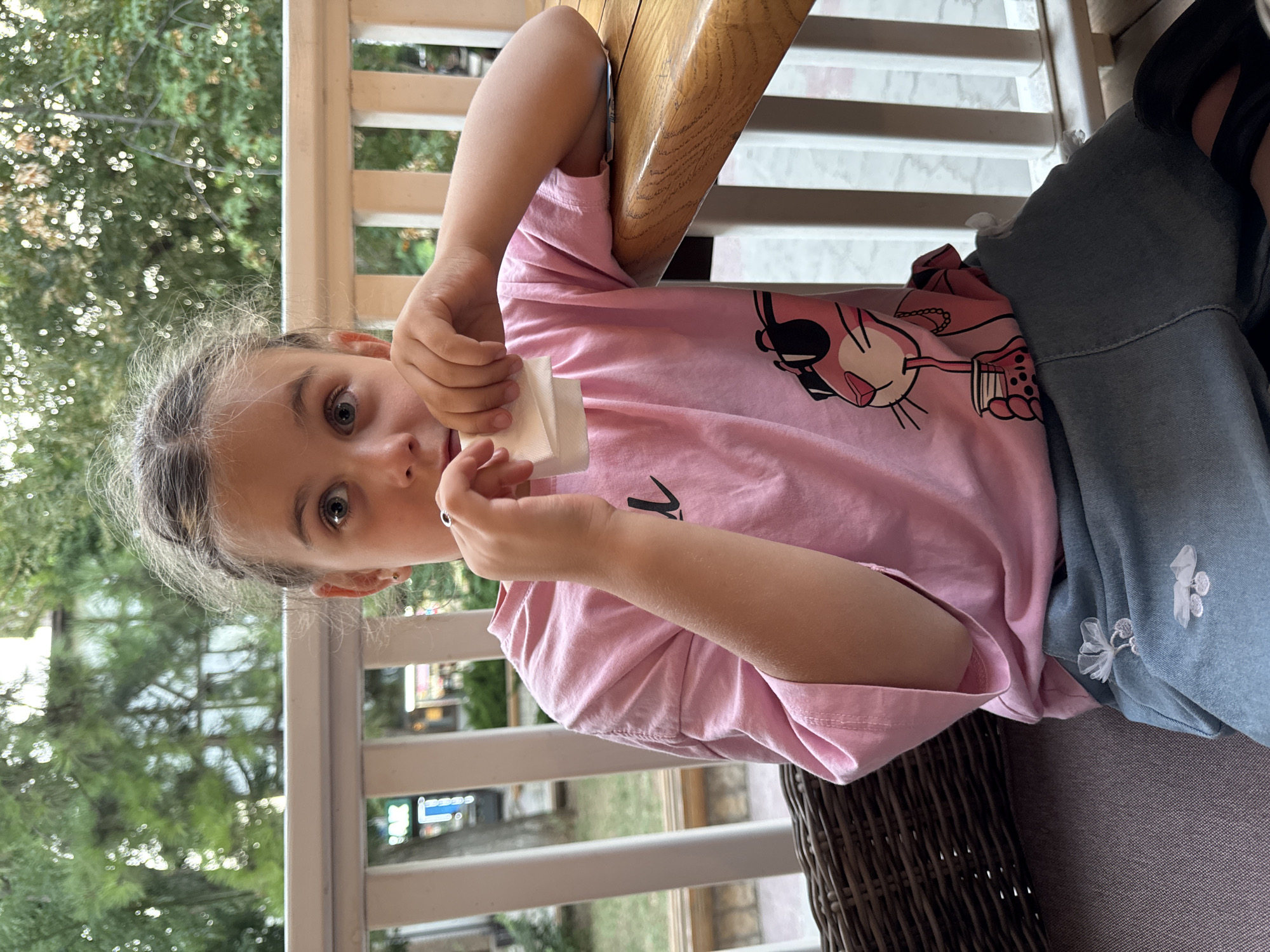 A young girl with braided hair is seated at an outdoor table on a porch in Yevpatoriya, Ukraine. She is wearing a pink t-shirt with a graphic of the Pink Panther and a denim skirt. She holds a white napkin to her mouth with both hands. The background shows a railing and greenery. The lighting suggests it is daytime.