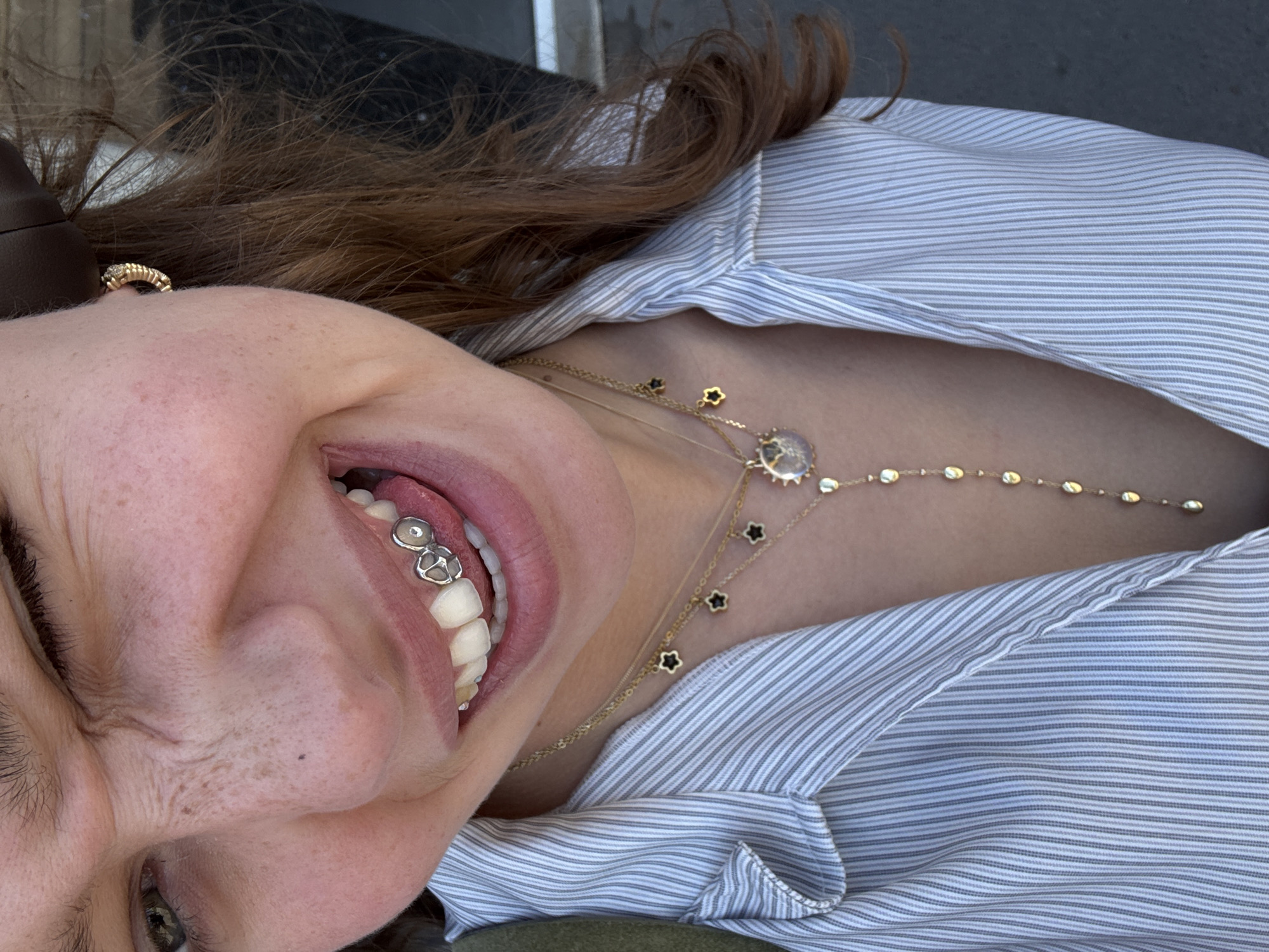 A close-up, off-kilter shot features a woman with auburn hair and freckled skin, wearing a striped shirt, lying back and smiling to reveal dental jewelry. She is wearing a gold necklace. A gold ring can be seen on her right temple. The location is undetermined, as is whether or not the photo was taken in Sofia, Bulgaria.