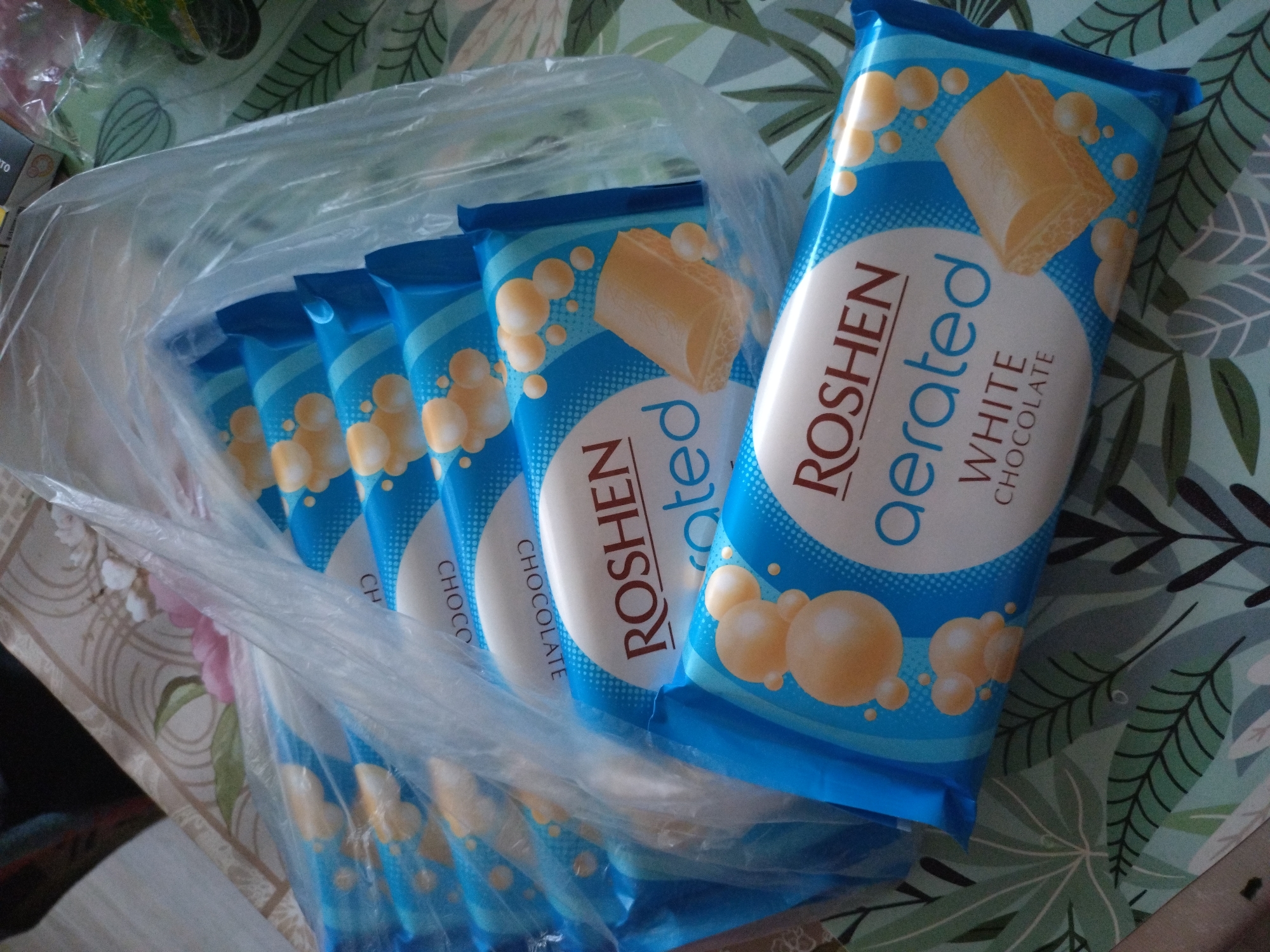 A slightly high-angle shot captures a cluster of Roshen Aerated White Chocolate bars, still partially encased in a clear plastic bag. The wrappers are primarily blue and white with brown lettering. An opened bag of the bars is on a table in a room. The table surface is patterned with stylized green and brown leaves, while other areas of the table are decorated with swirling patterns in off-white and light pink. The scene appears to be indoors, as evidenced by the tabletop and the presence of the plastic bag. The bars are the primary subject, arranged to showcase their packaging, with the plastic bag serving to group them. The bars are resting on a table, the table top appears to be in a room in Stara Zagora, Bulgaria.
