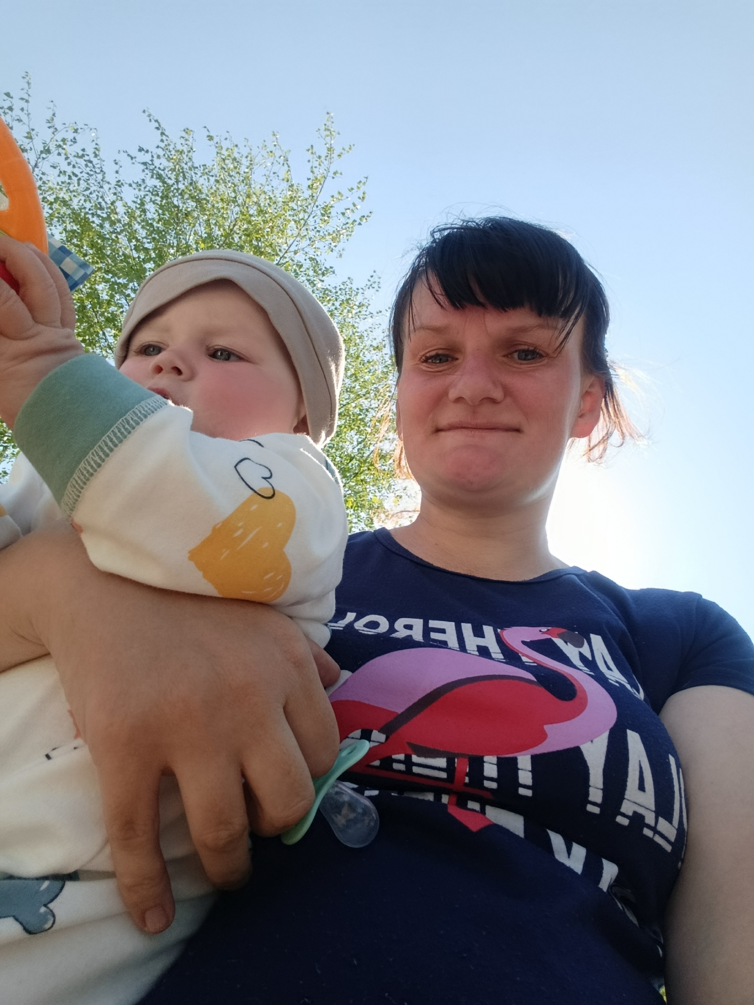 A woman holds a baby. The woman is wearing a navy blue t-shirt with a flamingo design and the baby is wearing a white long-sleeved shirt with an image of a yellow duck. The baby is wearing a beige beanie. The baby is holding a brightly colored toy. A pacifier is in the hand of the woman. A leafy tree is visible in the background against a blue sky. The location is outdoors and during the day. The city of Фоки, Russia, cannot be determined.