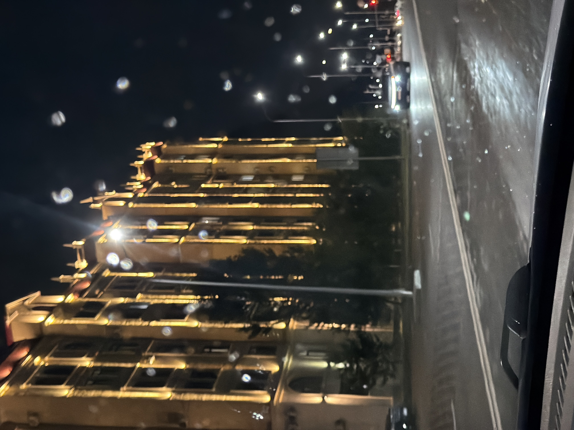 A night-time view from inside a vehicle shows a large, multi-story building with ornate architectural details illuminated by warm yellow lights. The building appears to be a hotel or apartment complex, with arched windows and decorative elements on its facade. In front of the building, trees are silhouetted against the lights. The street in the foreground is wet, suggesting it has recently rained, and reflects the surrounding lights. Several streetlights are visible, casting pools of light on the road. A few cars are driving on the street, their headlights visible. The sky is dark and appears clear, with some bokeh effects, possibly from raindrops on the vehicle's windshield. The overall scene depicts an urban environment at night in Astana, Kazakhstan, with a focus on the illuminated architecture and the wet street conditions.