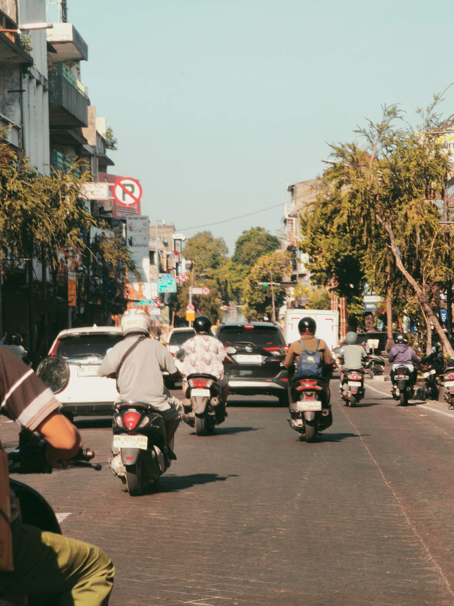 The image depicts a street scene in Denpasar, Indonesia, characterized by moderate traffic, primarily consisting of motorcycles. The perspective is from behind a motorcycle, looking forward down a wide, paved road. Buildings line both sides of the street, displaying a mix of architectural styles with balconies and trees providing some greenery. Several traffic signs are visible, including a red and white "no parking" sign.
In the foreground, partially obscured, is the back of a person wearing a striped shirt and olive-green pants, seated on a motorcycle. Following this are multiple motorcycles with riders, some carrying passengers. One prominent white car is visible in the middle ground, and several other vehicles are present. The road surface appears to be asphalt with a textured pattern. The sky is a clear, pale blue. The overall atmosphere suggests a typical day in a bustling urban environment.