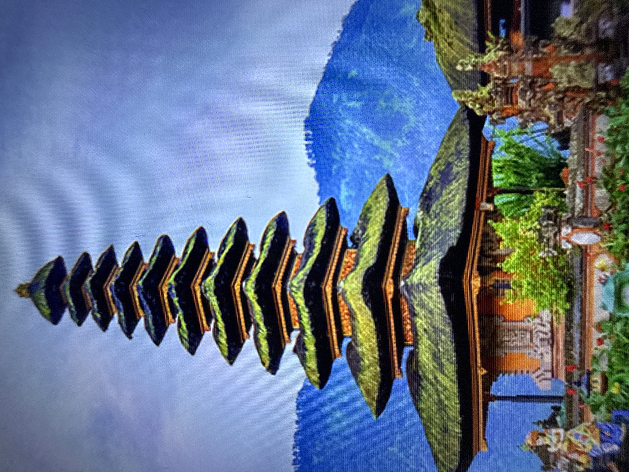The image captures a Pura Ulun Danu Bratan temple in Bali, Indonesia, set against a backdrop of lush green mountains under a clear blue sky. The temple features multiple tiers of tiered roofs, characteristic of Balinese architecture, with the roofs covered in vibrant green moss. The foreground shows intricate stone carvings and statues, along with colorful floral arrangements. The overall scene evokes a sense of serenity and spiritual grandeur, highlighting the temple's scenic location and architectural beauty.