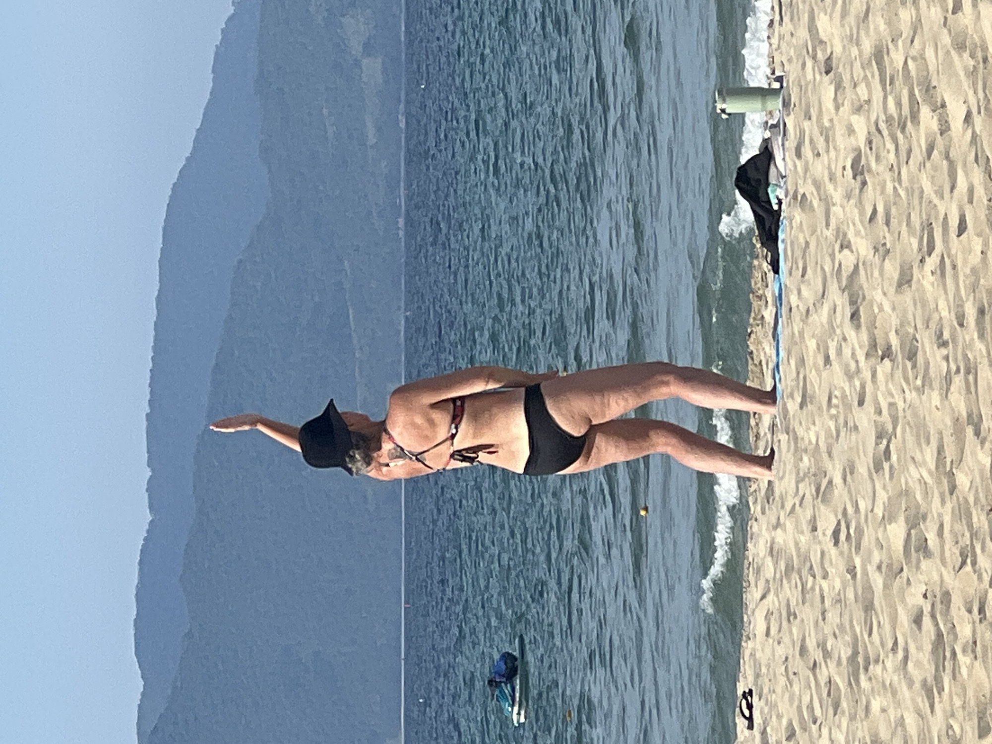 The image shows a woman in a black bikini and a black hat on a sandy beach. She is leaning back with her left arm extended, as if in mid-movement during a yoga pose or dance. The background consists of the blue ocean with gentle waves, and distant, hazy mountains. The sun appears to be high in the sky, suggesting it is daytime. Some personal items, including a light green water bottle and a dark bag, are visible on the sand near the water's edge. A few small boats or kayaks are floating in the water further out. The overall scene is tranquil and relaxed, typical of a beach environment. The image is taken in Đà Nẵng, Vietnam.