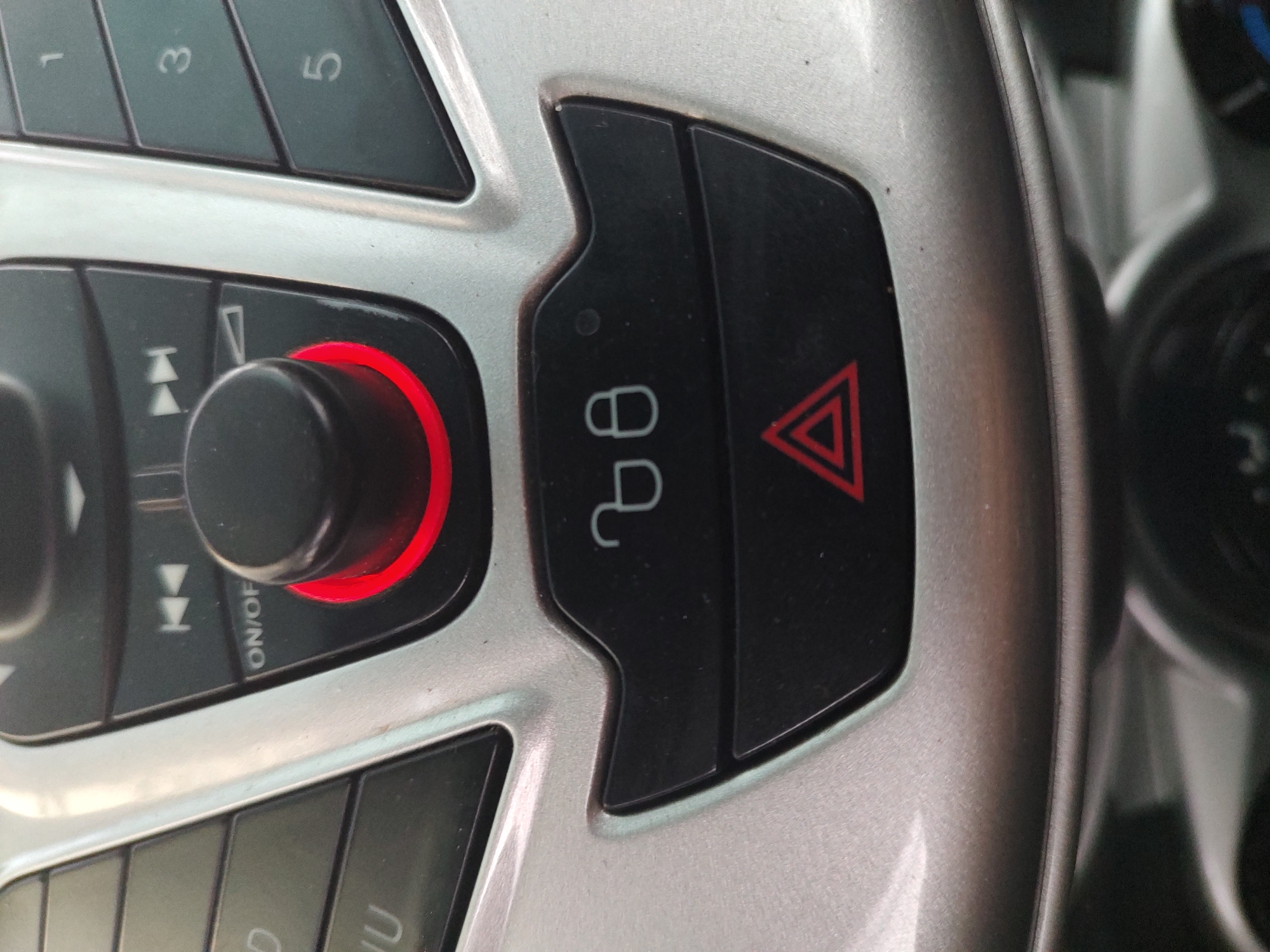 A close-up, eye-level shot shows the center console of a car's dashboard, specifically focusing on the audio controls and hazard light button. The console features silver plastic surrounding black buttons and a central black knob with a red illuminated ring. To the left of the knob are directional buttons, one labeled "ON/OFF." To the right are numbered buttons: 1, 3, and 5 are visible. Below the audio controls, on a black surface, are two white icons: a lock symbol and an unlock symbol. Directly beneath these is the hazard light button, a black square with a red equilateral triangle inside. The background is blurred, but suggests the interior of a vehicle. There are no people or animals visible. The image appears to be taken in the city of Tervel, Bulgaria, based on the metadata, but this context is not visually evident in the photograph itself.