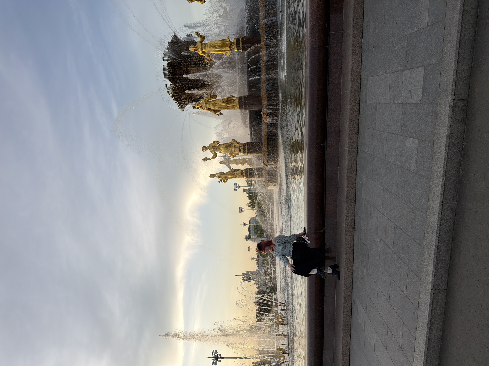 A vertically oriented medium outdoor shot depicts the Friendship of Nations Fountain in Moscow, Russia. The fountain is composed of several tiers. On the top tier, sixteen gold statues of women circle the fountain basin. Water is cascading from the fountain into the basin in a series of streams and arcs. A woman in a light-colored shirt and black skirt is seated on the edge of the fountain. The woman is facing the fountain, and she is holding her phone. In the background, there are several tall buildings and street lights. The sky is pale blue. The foreground is a gray stone tiled walkway.