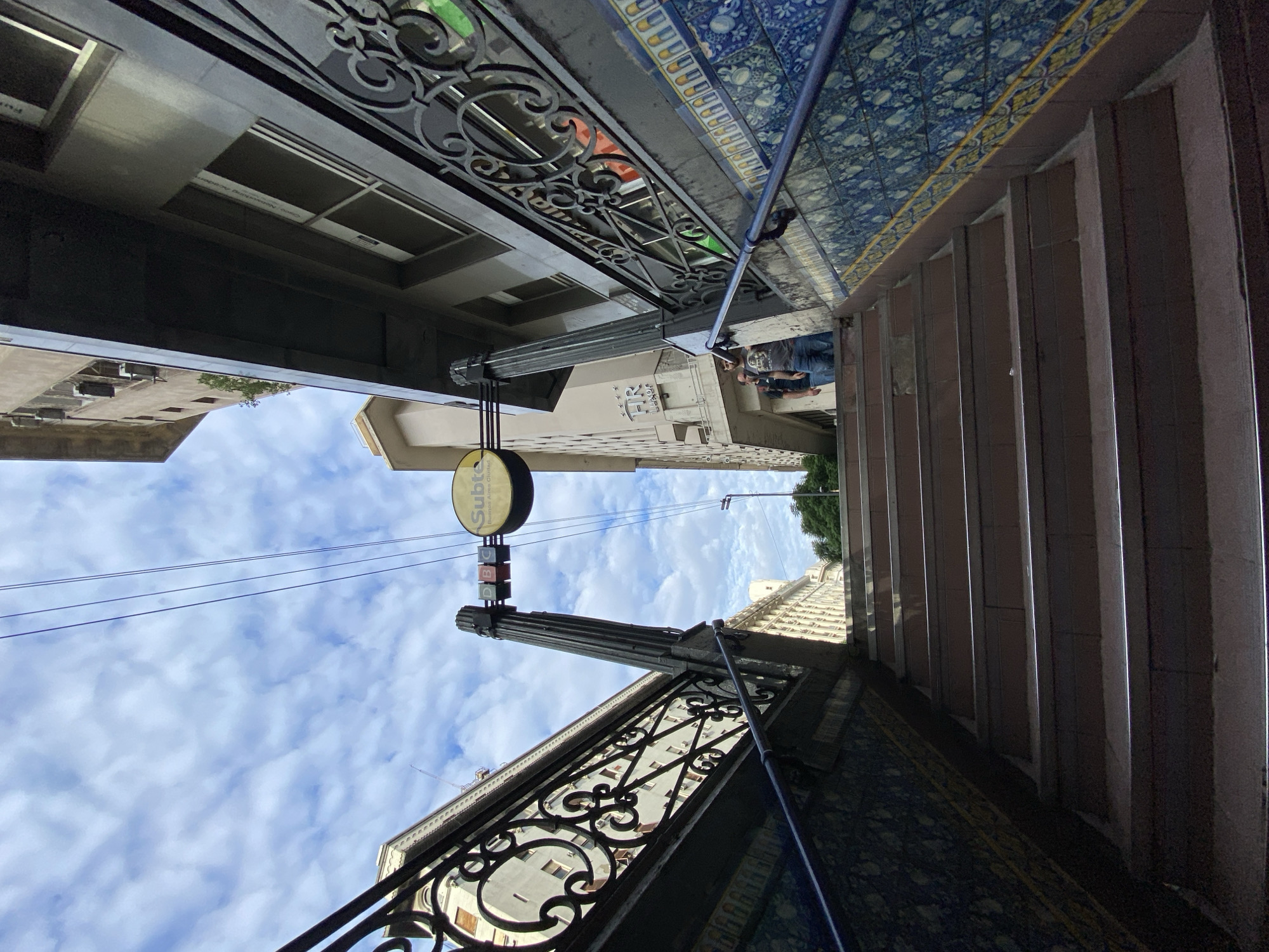 The image depicts a view of a "Subte" (subway) entrance, located in Buenos Aires, Argentina. The perspective is from below, looking upwards through the entrance frame. An ornate metal railing with scrollwork designs is visible on both sides of the frame. Above the railing on one side, decorative blue and yellow tiles line the ceiling. Steps lead upwards from the entrance. A round "Subte" sign is mounted on a fluted column. Above the sign, power lines stretch across the sky filled with clouds. Buildings line the street in the background. A man wearing a T-shirt is standing near the steps.