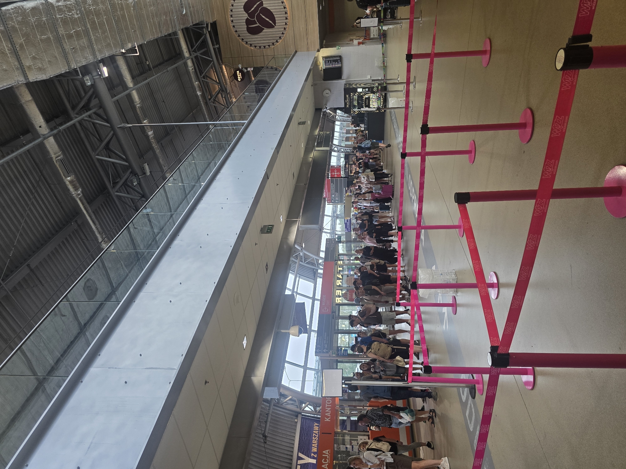 The image depicts a queue of people at what appears to be an airport or train station in Nowy Dwór Mazowiecki, Poland, likely Warsaw Modlin Airport, given the "WIZZ" branding on the queue barriers. The scene is indoors, with a high ceiling featuring exposed industrial ductwork and lighting. A second-level walkway with a glass balustrade overlooks the main area. People are standing in line, many with luggage, suggesting they are waiting for a flight or train. They are positioned behind pink stanchions with retractable barriers that bear the "WIZZ" logo. In the background, there are signs indicating "Kantor" (currency exchange) and what appear to be departure gates. A Costa Coffee sign is visible on the upper right wall, and a large graphic of coffee beans is also present. The overall environment is brightly lit and functional, typical of a transportation hub.