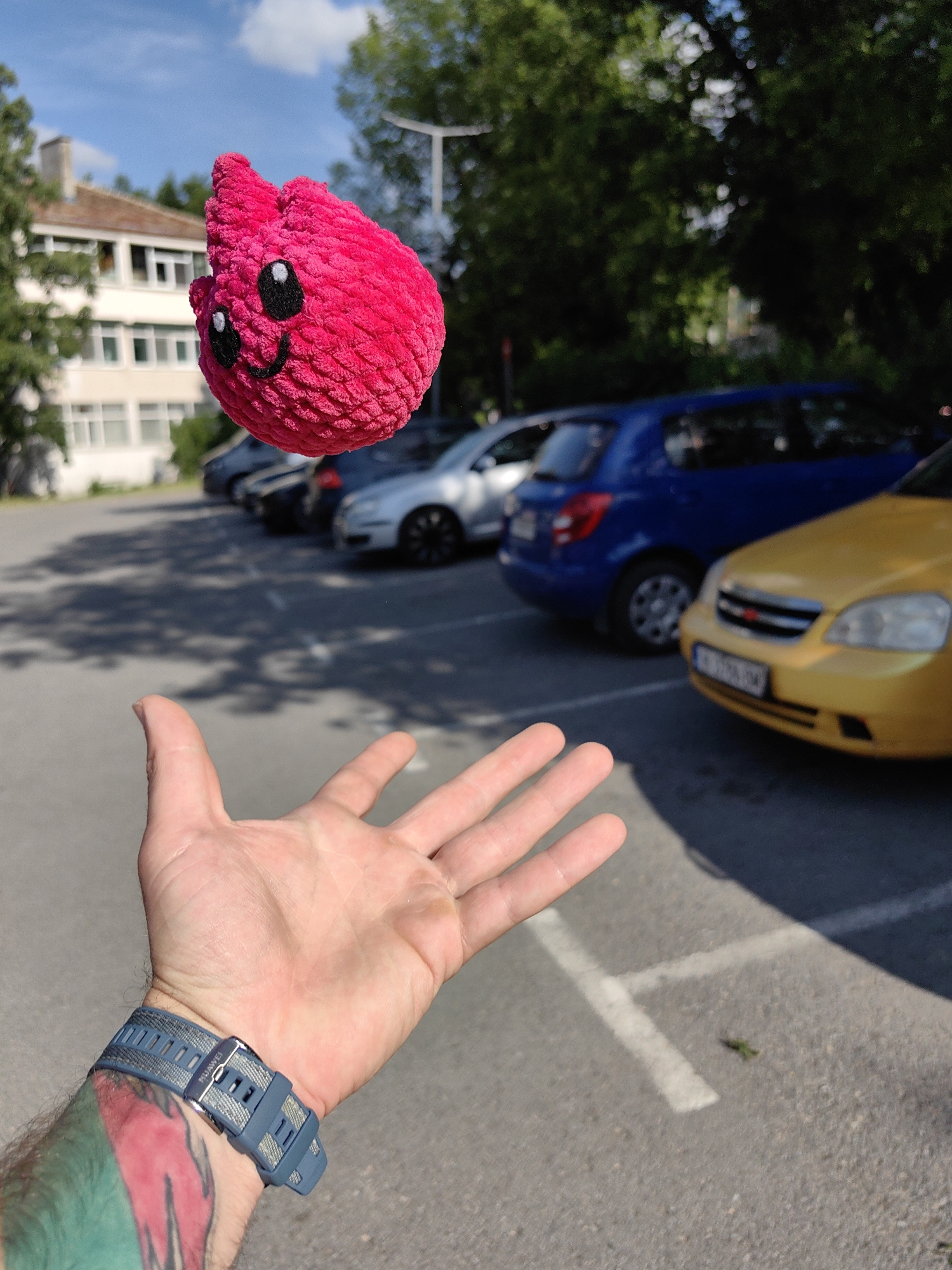 A hand with a blue smart watch is reaching towards a small, fuzzy, pink, anthropomorphic toy that is floating in the air, with a parking lot and building in the background on a sunny day. A yellow, blue, silver, and black vehicle can be seen, parked on the lot. The location is identified as the city of Shumen, Bulgaria.