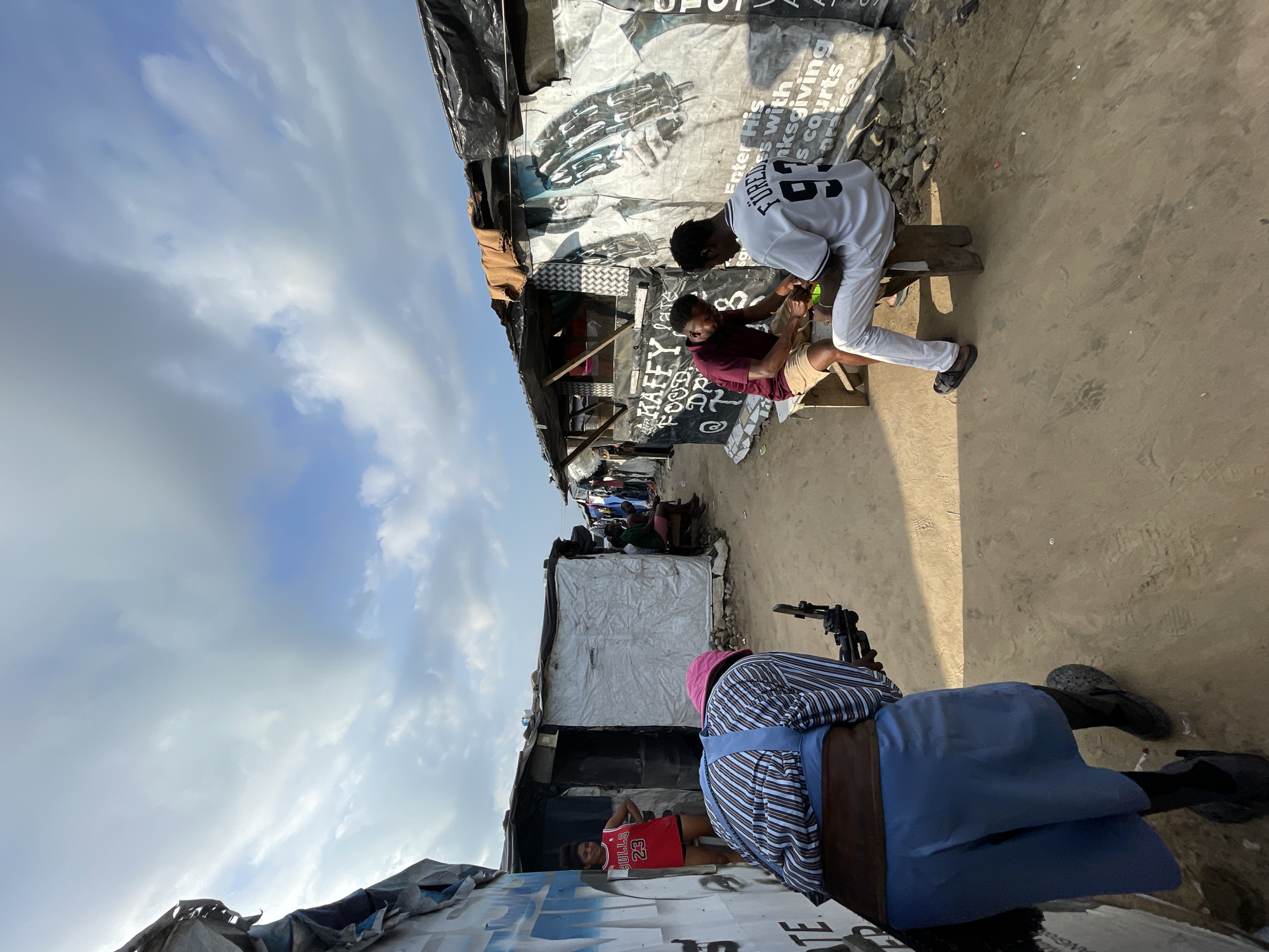 This image depicts a scene in Iranla, Nigeria. A person wearing a pink hat, a striped shirt, a brown belt, and a blue skirt is holding a camera, seemingly filming. In the background, two men are seated on a bench in front of a makeshift stall with posters on the wall. The stall appears to be selling food and drinks. A young woman wearing a red Bulls basketball jersey stands near another stall to the left. The environment is characterized by informal structures constructed from corrugated metal and plastic sheeting, common in informal settlements. The ground is sandy. The sky is partly cloudy.