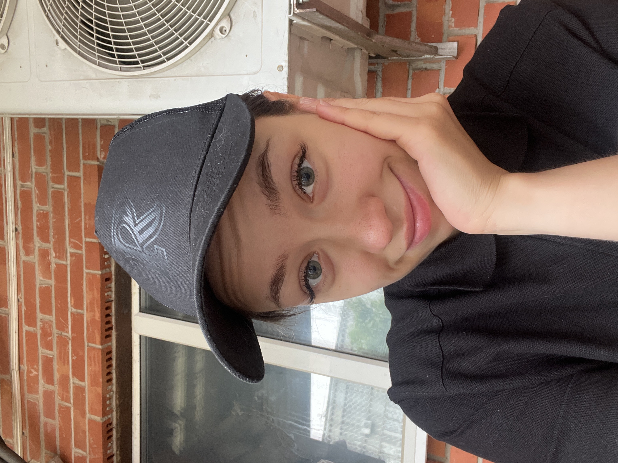 In this image, a young woman wearing a black cap and black polo shirt is posing for a selfie outdoors. Her right hand is supporting her chin, and she is looking at the camera with a slight smile. Behind her, a brick wall and an air conditioning unit are visible, suggesting an urban or residential setting, possibly in Chaikovsky, Russia, given the context. The lighting indicates it is daytime.
