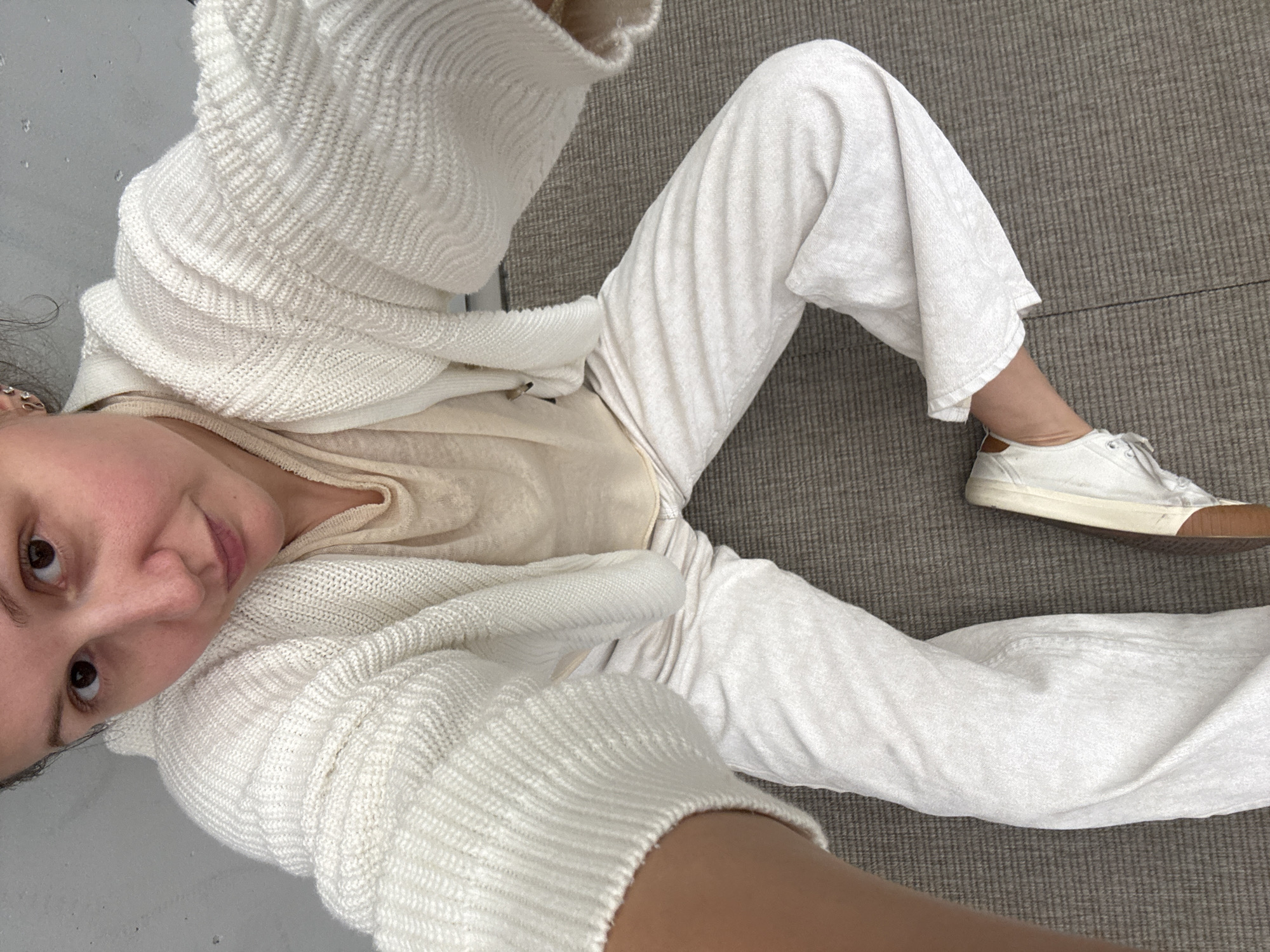 A woman is lying on a gray carpet, wearing white pants, a white shirt, and a white cardigan. She has on white sneakers with a brown sole. The photo appears to be a selfie. There is no evidence in the image that suggests the location is Dubai, United Arab Emirates.