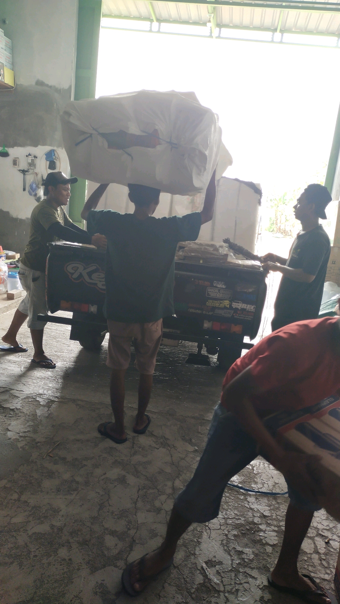 In the city of Pandanarum, Indonesia, four individuals are engaged in the process of loading a large, white, tarp-wrapped object onto the back of a dark-colored truck. The truck has text and logos visible on its side. One person, wearing a green shirt and a dark cap, is on the left, assisting in the loading. Another person, wearing a dark green t-shirt and light-colored shorts, is carrying the object on their head. A third person, wearing a dark t-shirt and a dark cap, is standing on the right, observing the loading process. A fourth person, wearing a red shirt and dark shorts, is in the foreground, partially obscured and also observing. The environment appears to be a warehouse or open-sided structure with a concrete floor. The lighting suggests it is daytime, with bright light coming from an opening or window.