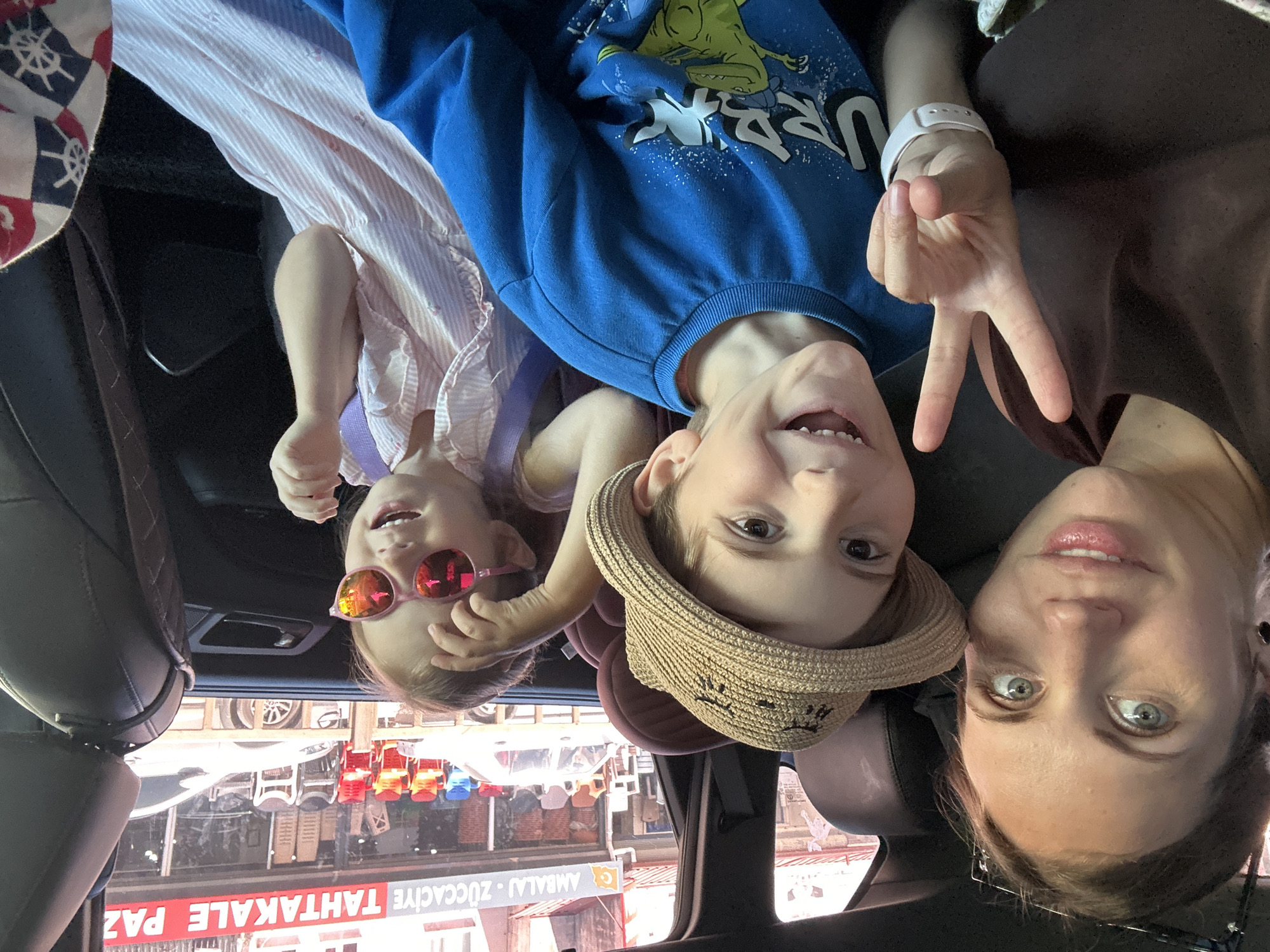 The image depicts a woman and two children in what appears to be the back seat of a vehicle. The woman is on the right side of the frame, with her face clearly visible and angled towards the camera. A younger child, wearing a straw hat, is positioned to her left, holding up a peace sign. Another child, wearing pink sunglasses, is on the left side of the frame, next to the window. The vehicle is parked outside a shop. A reflection visible in the window of the shop says "ZOCCACIYE - AMBALAJ - ZOCCACIYE - TAHTAKALE - PAZ". Based on the location this shop is in the city of Fethiye, Turkey.