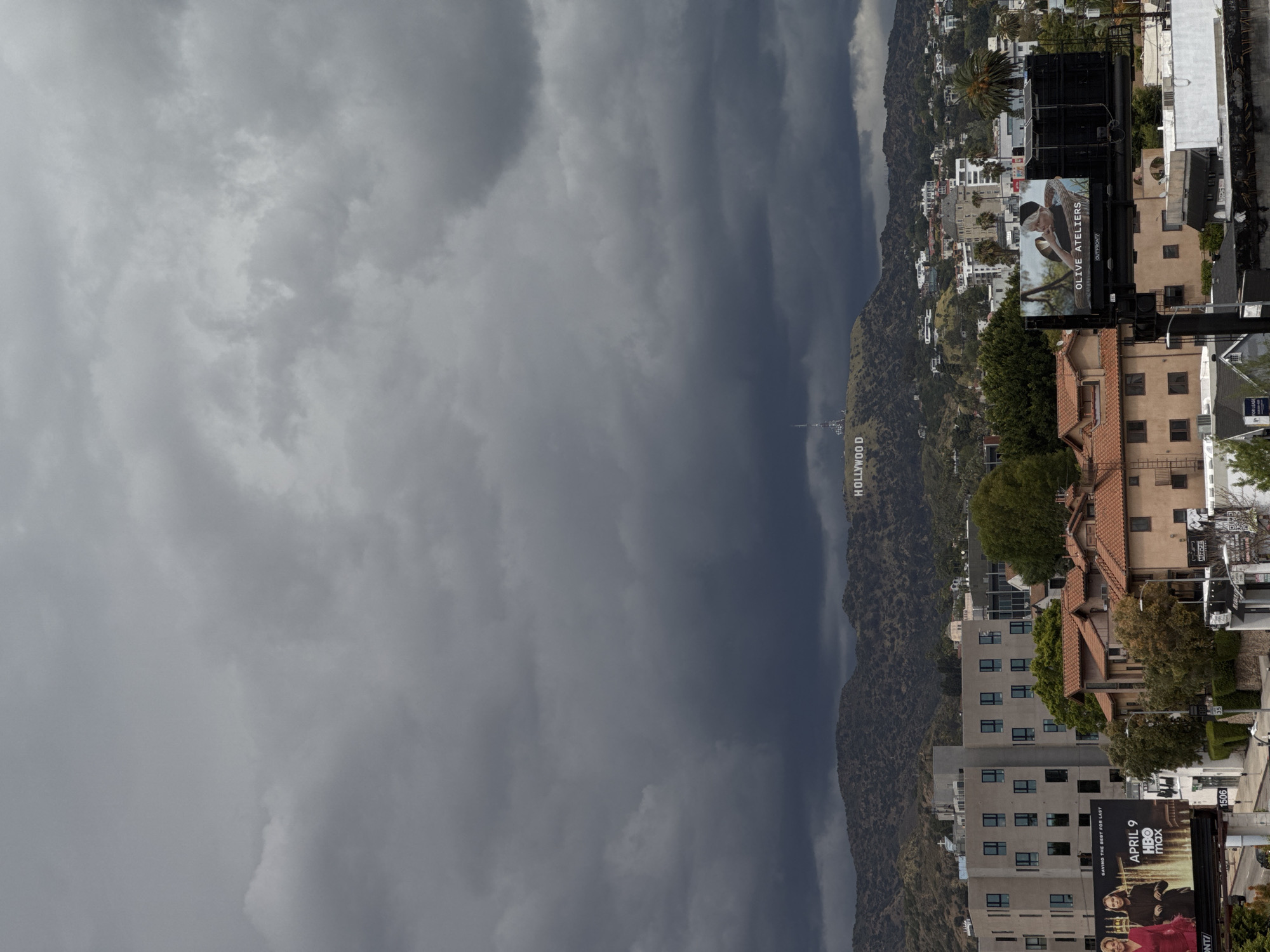 Here is a detailed description of the image:
The image showcases the iconic Hollywood Sign nestled into the hills, with a cloudy, overcast sky dominating the background. The sign is located on the hills and is viewed from a high-rise, with the city of Los Angeles sprawling in the foreground.
Several buildings and billboards are also in the frame. The billboards promote different media, with the "OLIVE ATELIERS" billboard and the "APRIL 9 HBO max" billboard prominently displayed. The overcast sky suggests a somewhat gloomy or potentially rainy day in Hollywood.