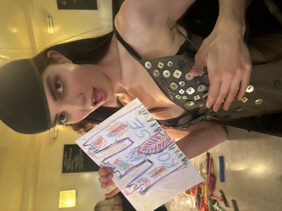 A woman in a dark top with iridescent buttons poses at an angle, holding up a child's crayon drawing. The drawing, on white paper, depicts colorful shapes resembling buildings and a feather. The words "ROSE" are scrawled beside the feather. A black cap covers the woman's hair. A table with crayons scattered across it is visible at the bottom of the frame.